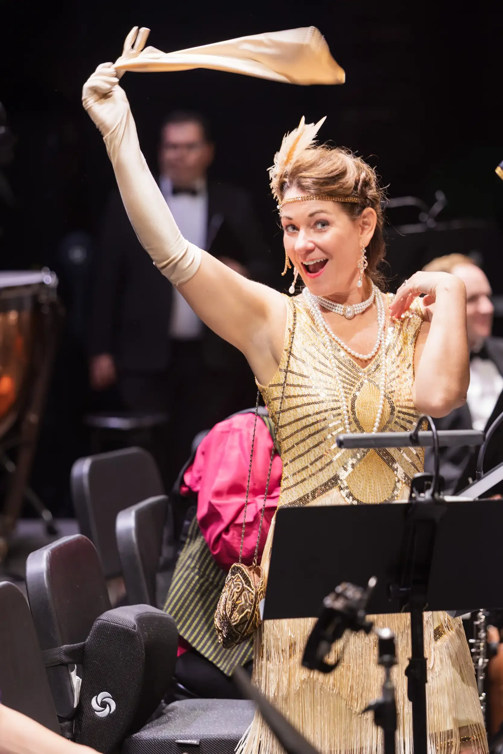 A woman dressed as a vintage flapper in a gold and black dress, wearing long gloves, pearls, and a feather headband, striking a playful pose in a dance hall or theater setting.