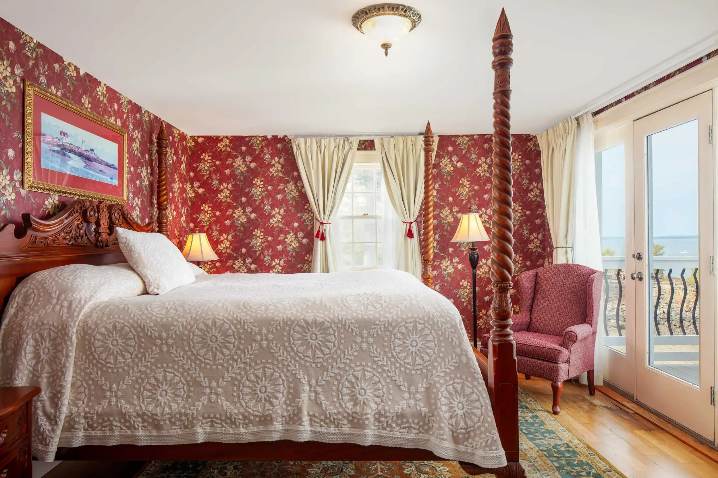 A bedroom with a four-poster bed with intricately carved wooden posts, a white quilt with a floral pattern, and pillows. The wall behind the bed is covered with red floral wallpaper. There are two table lamps on bedside tables, and cream curtains are