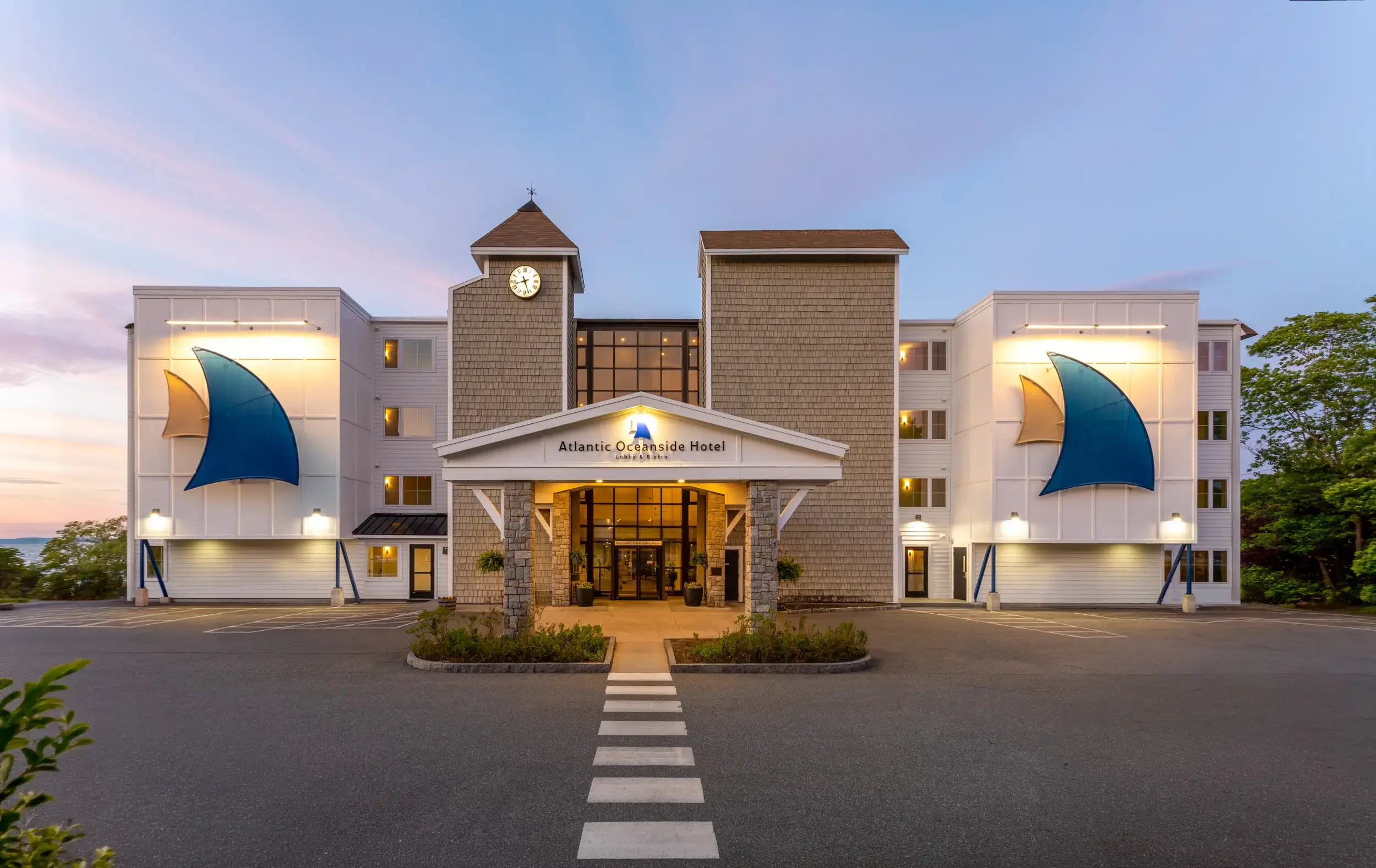 Front view of Atlantic Oceanside Hotel with a driveway and striped crosswalk leading to the entrance, featuring a clock tower and blue sails as architectural accents, during sunset with a clear sky and some trees in the background.