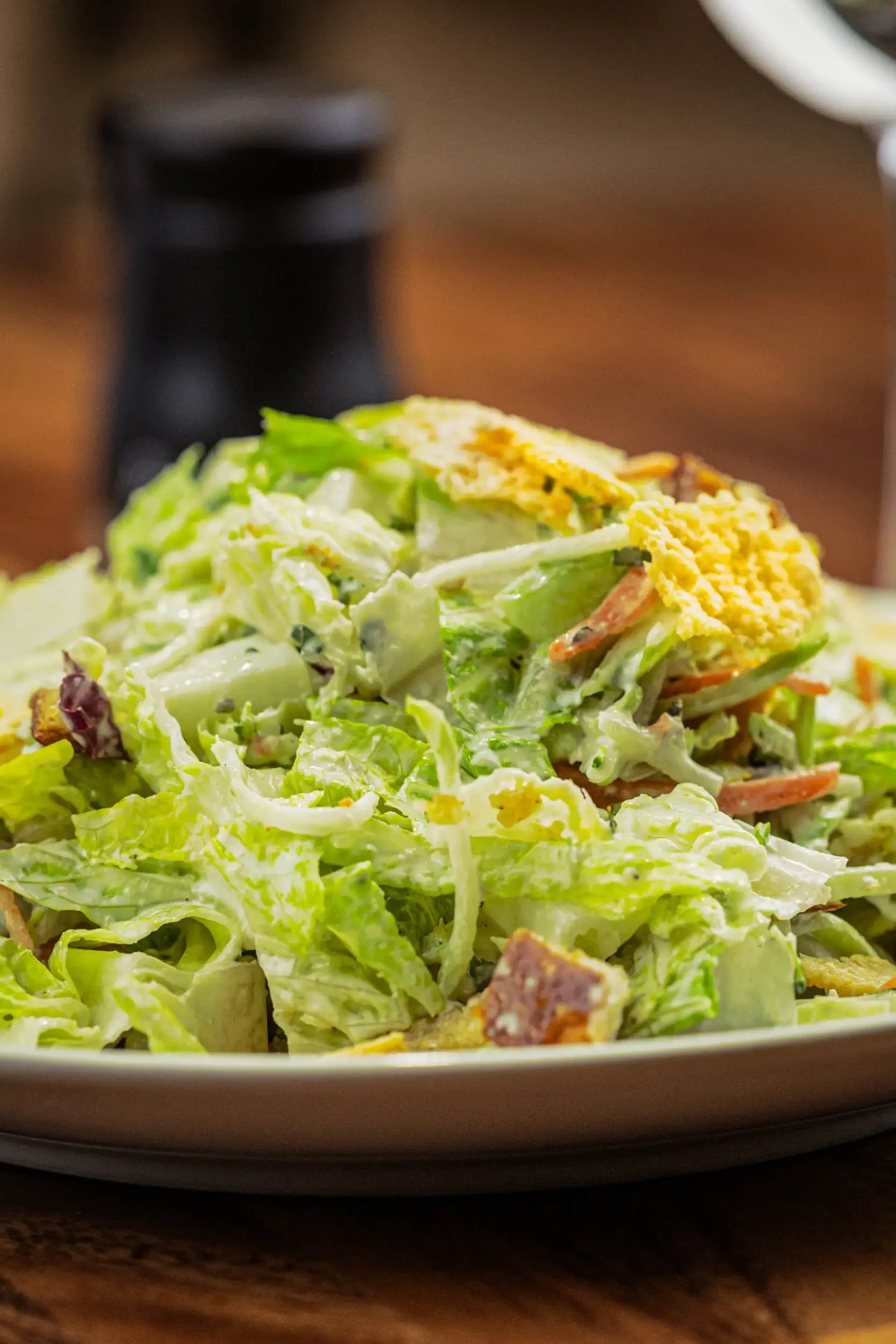 A close-up of a plate of Caesar salad topped with shredded cheese and croutons, with a blurred dark container in the background.