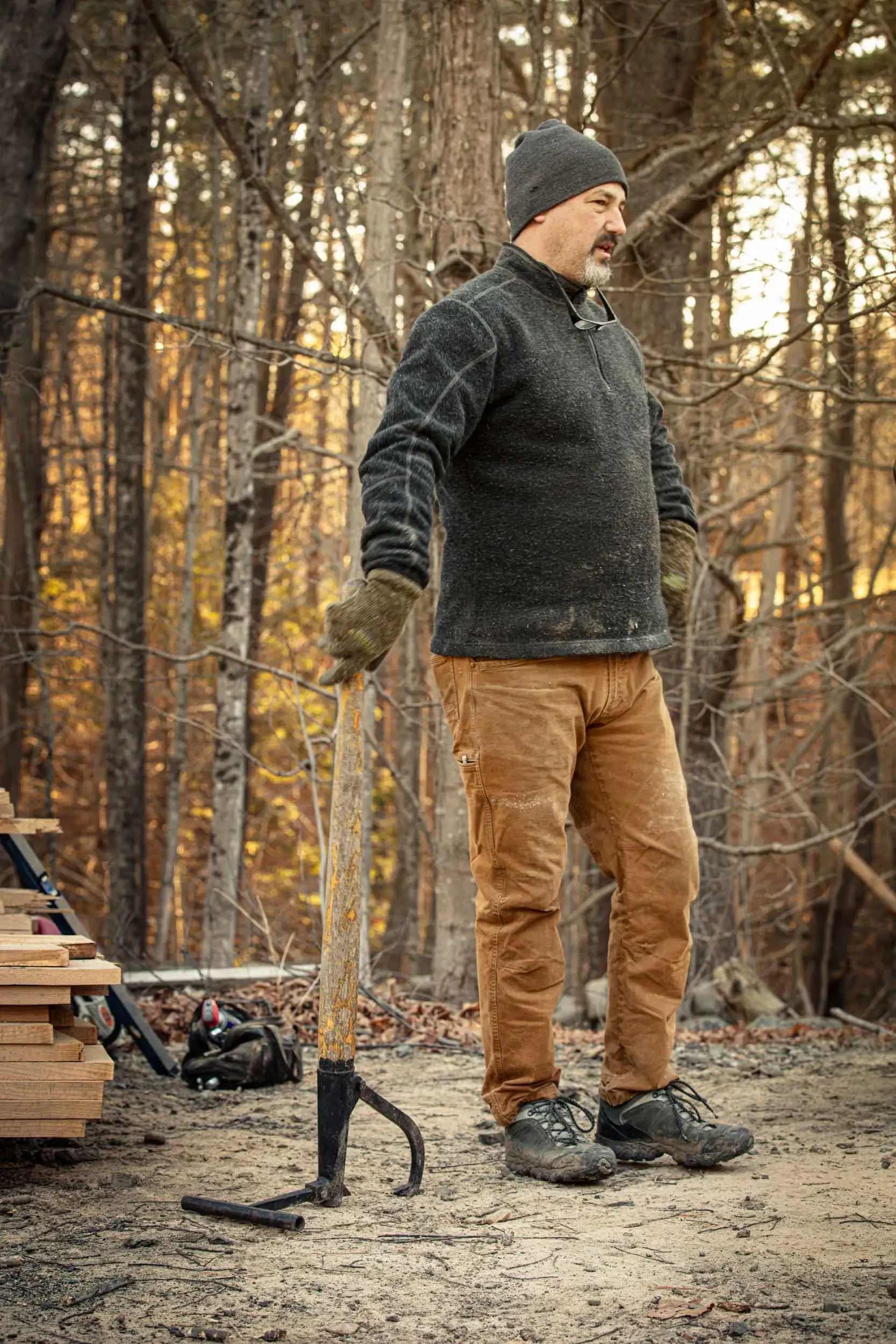 A man wearing a gray beanie, dark gray fleece jacket, brown work pants, and hiking boots standing in a wooded area during fall. He appears to be holding a tool and is surrounded by trees with autumn leaves. There are tools, wooden planks, and a backp