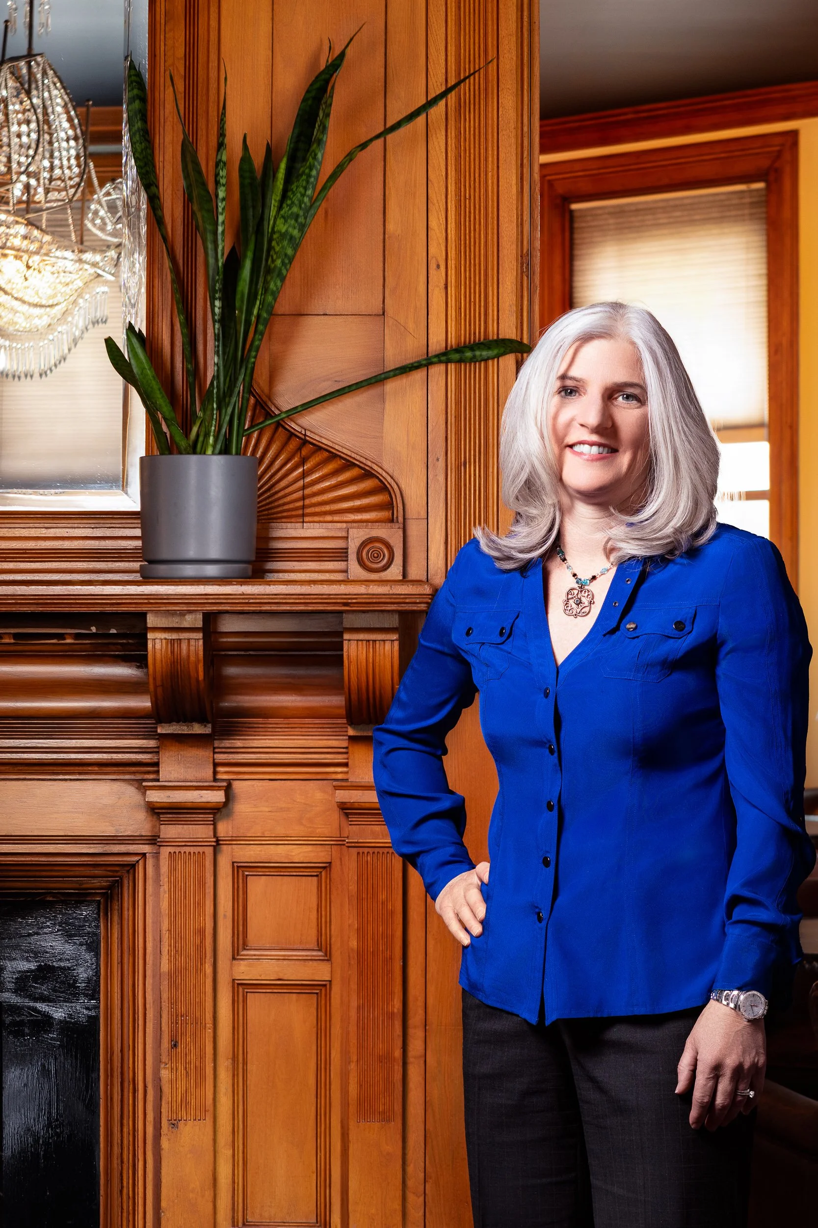 A woman with long gray hair standing indoors near a wooden wall with a large green potted plant nearby, wearing a blue shirt and smiling.