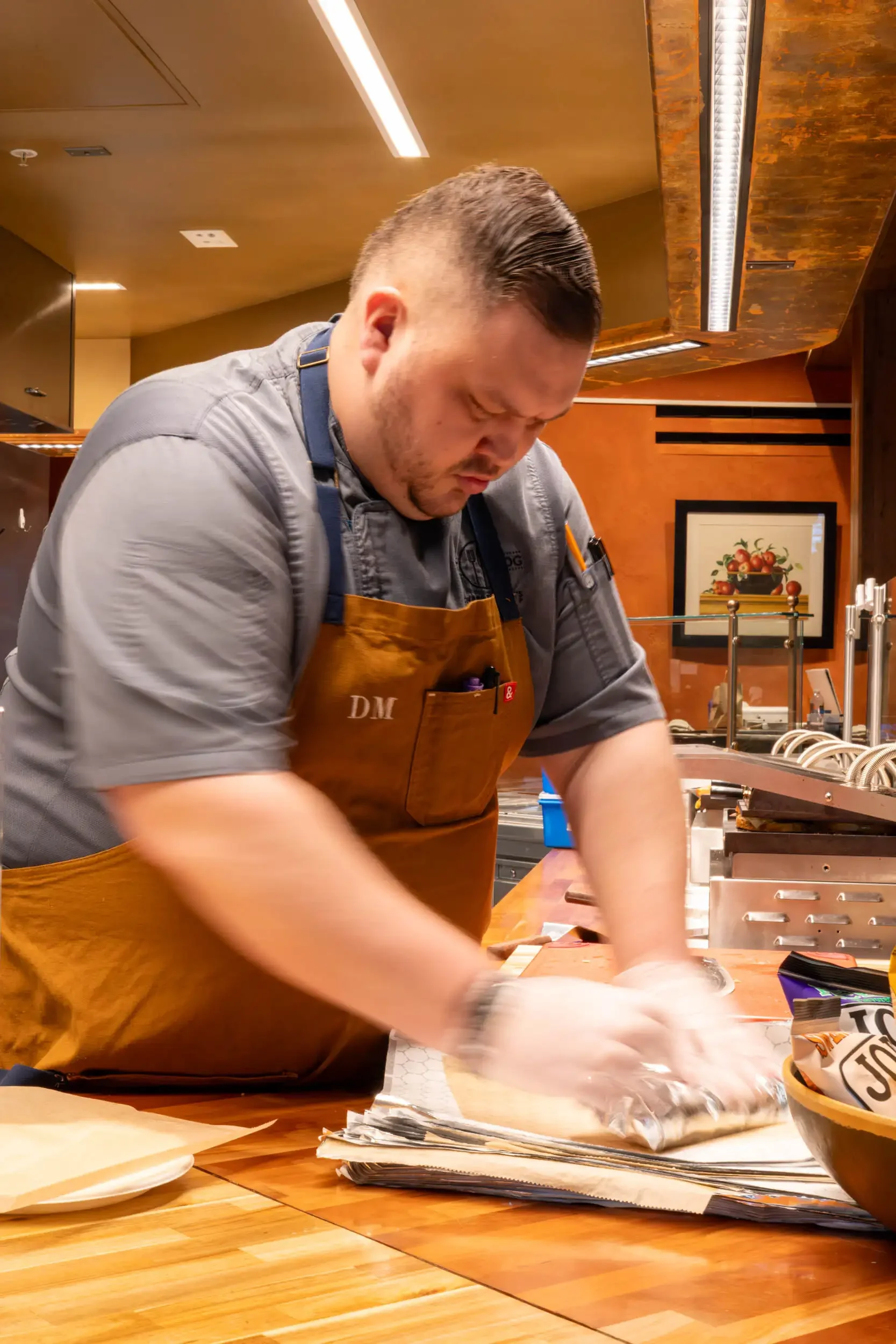 A man working in a kitchen, wearing an apron and gray shirt, with grooming on his hair, surrounded by kitchen and baking supplies.