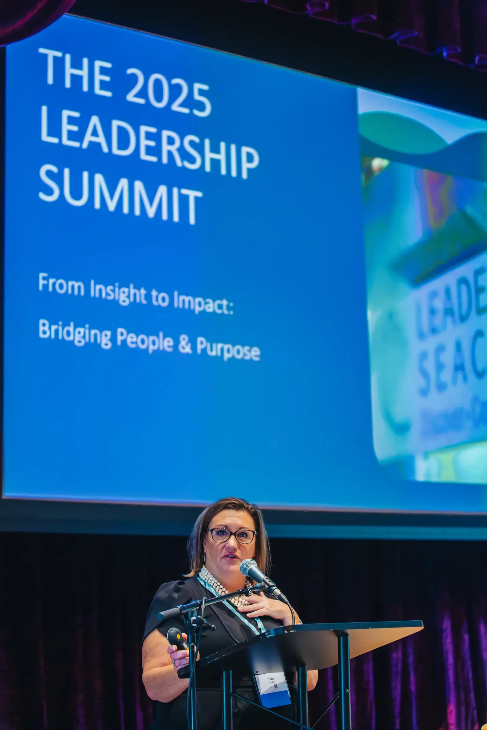 A woman with glasses, wearing a black dress with a pearl necklace, speaking at a podium with a microphone during the 2025 Leadership Summit. A large screen behind her displays the event title and theme.