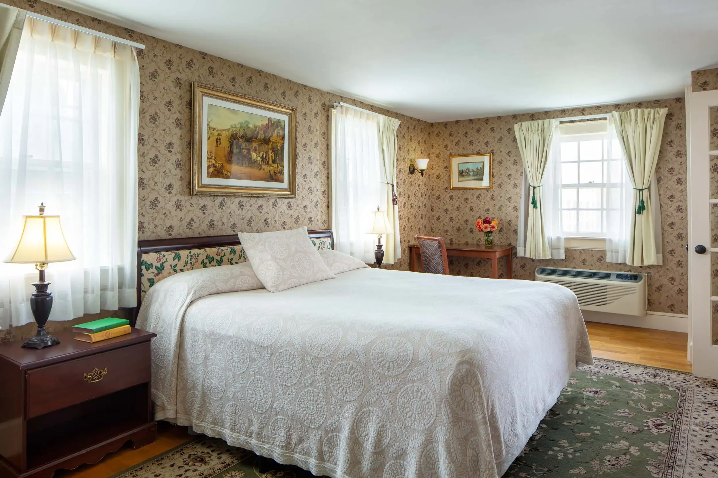 A cozy bedroom with a bed covered in a white quilt, nightstands with lamps, framed artwork on floral wallpaper, and large windows with cream curtains, sunlight streaming in.
