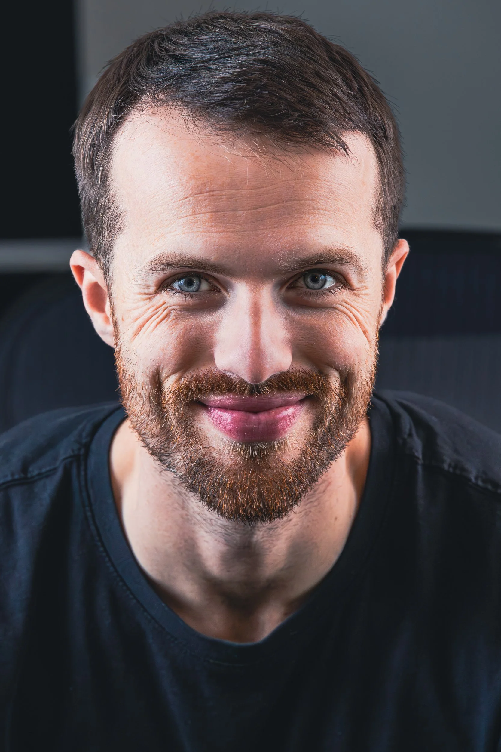 Close-up of a smiling man with a beard and blue eyes sitting indoors.