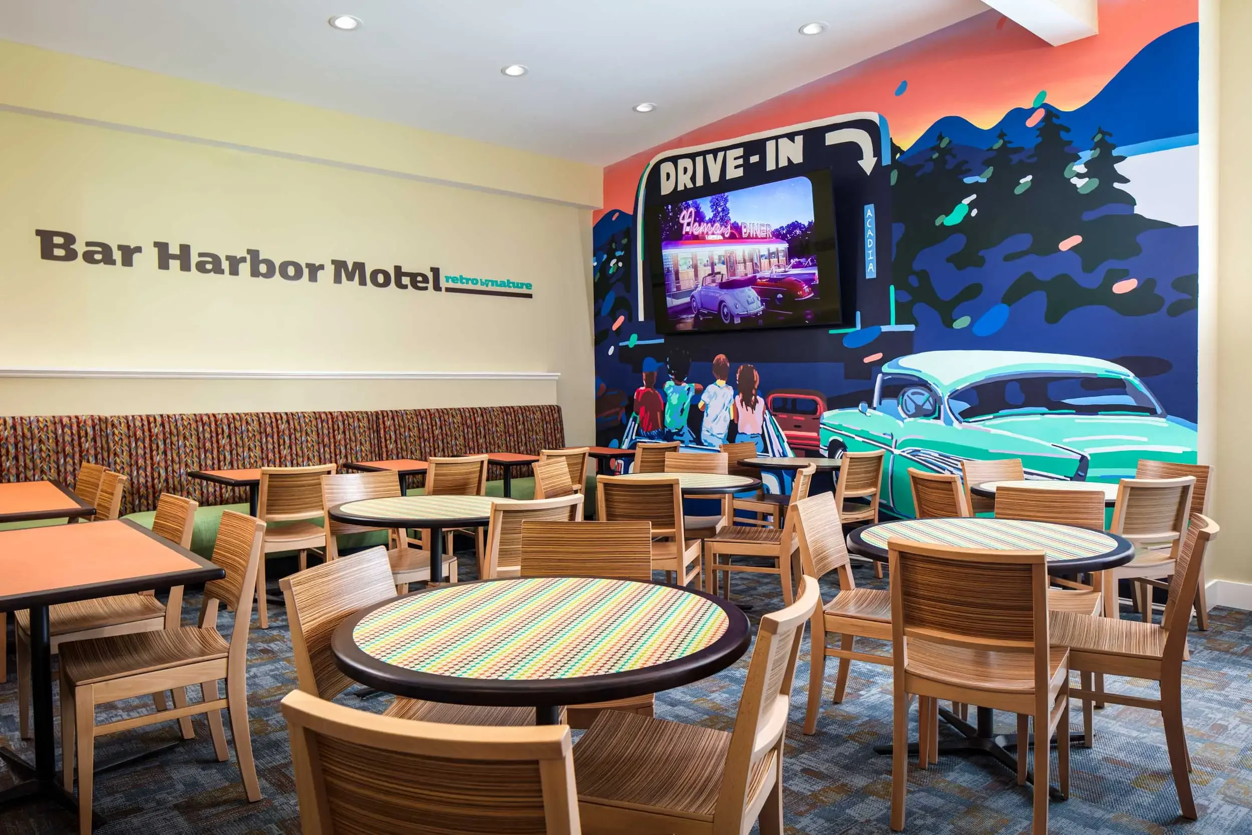 Interior of a diner with vintage style, wooden chairs and round tables, a colorful mural of mountains and cars, and a digital screen displaying a neon sign and classic cars.