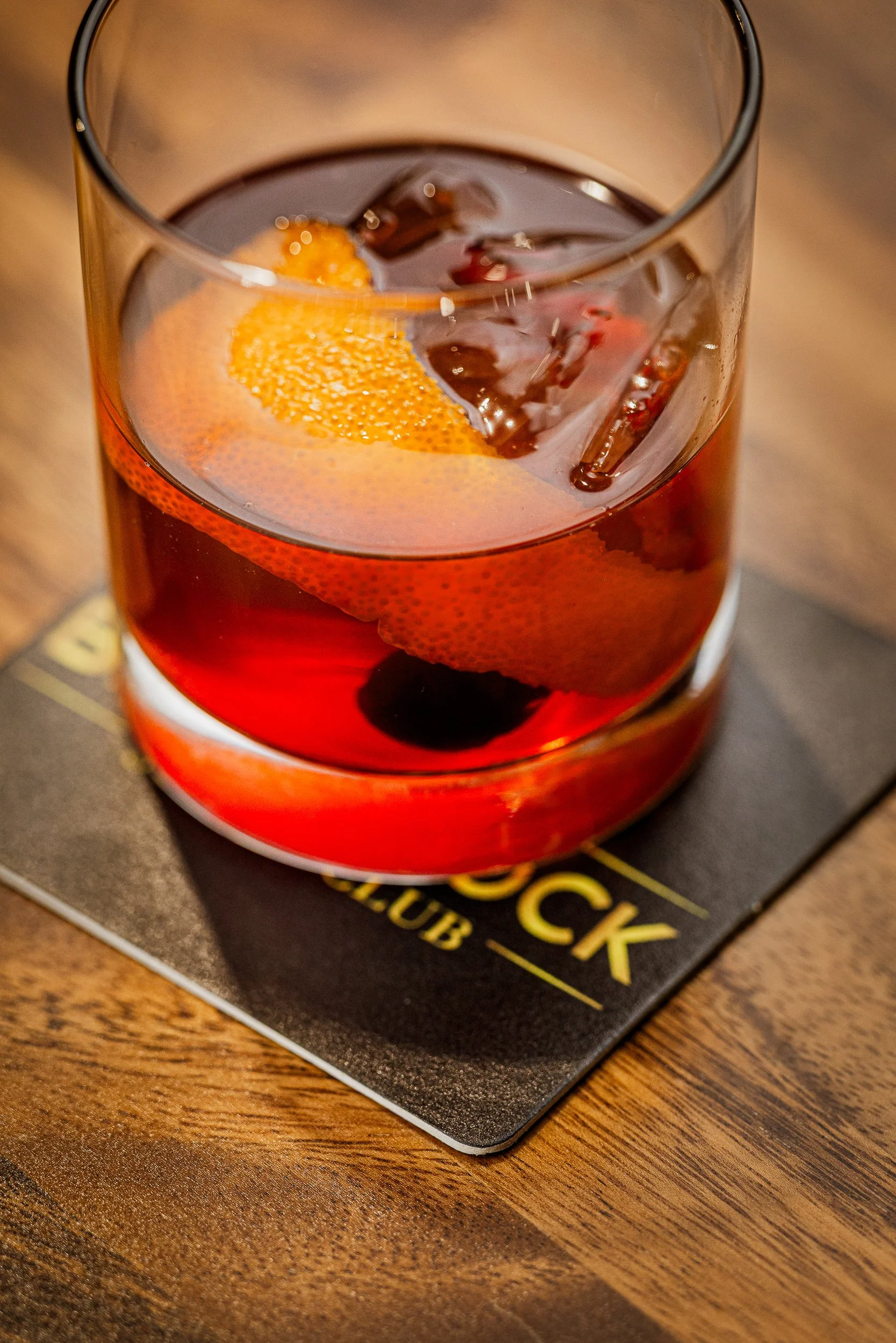 A glass of dark-colored cocktail with ice and an orange slice, placed on a black drink coaster on a wooden surface.