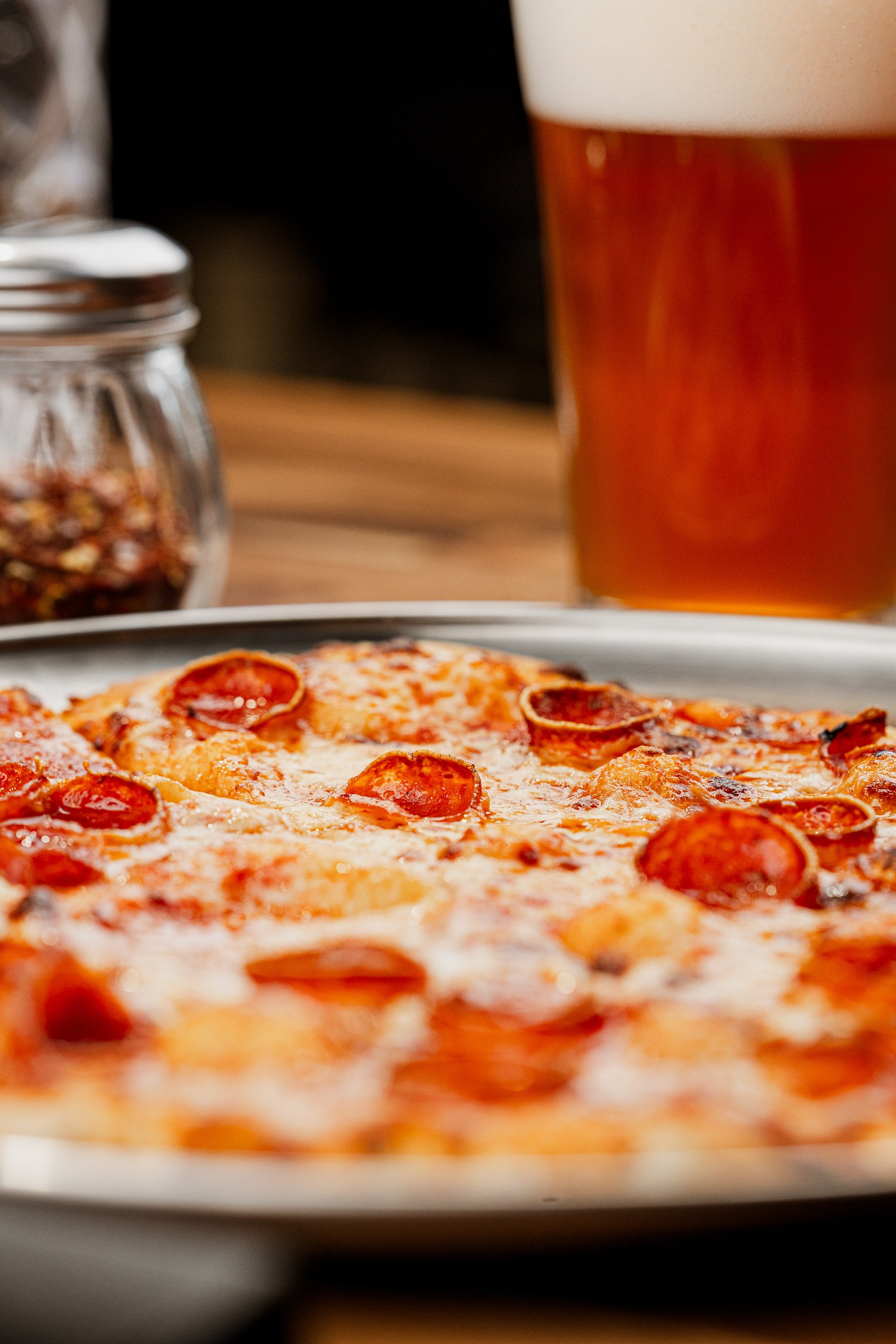 Close-up of a pepperoni pizza on a white plate with a mug of beer in the background and a jar of red pepper flakes on the side.