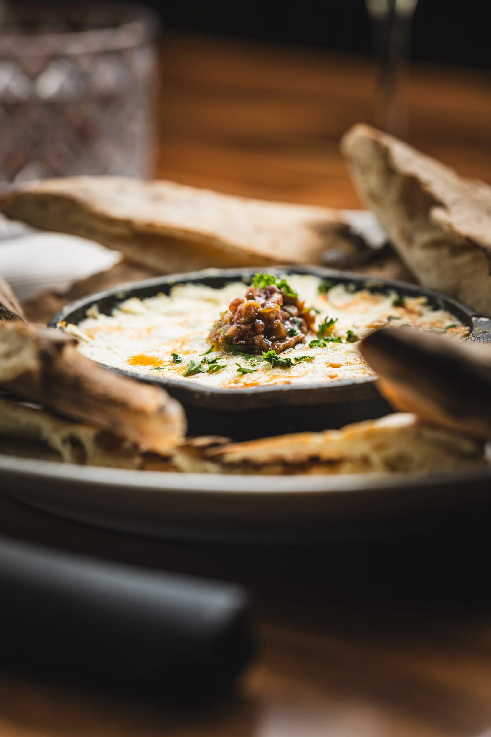 Baked cheese dip with a meat topping garnished with chopped herbs, surrounded by toasted bread slices on a dark plate.