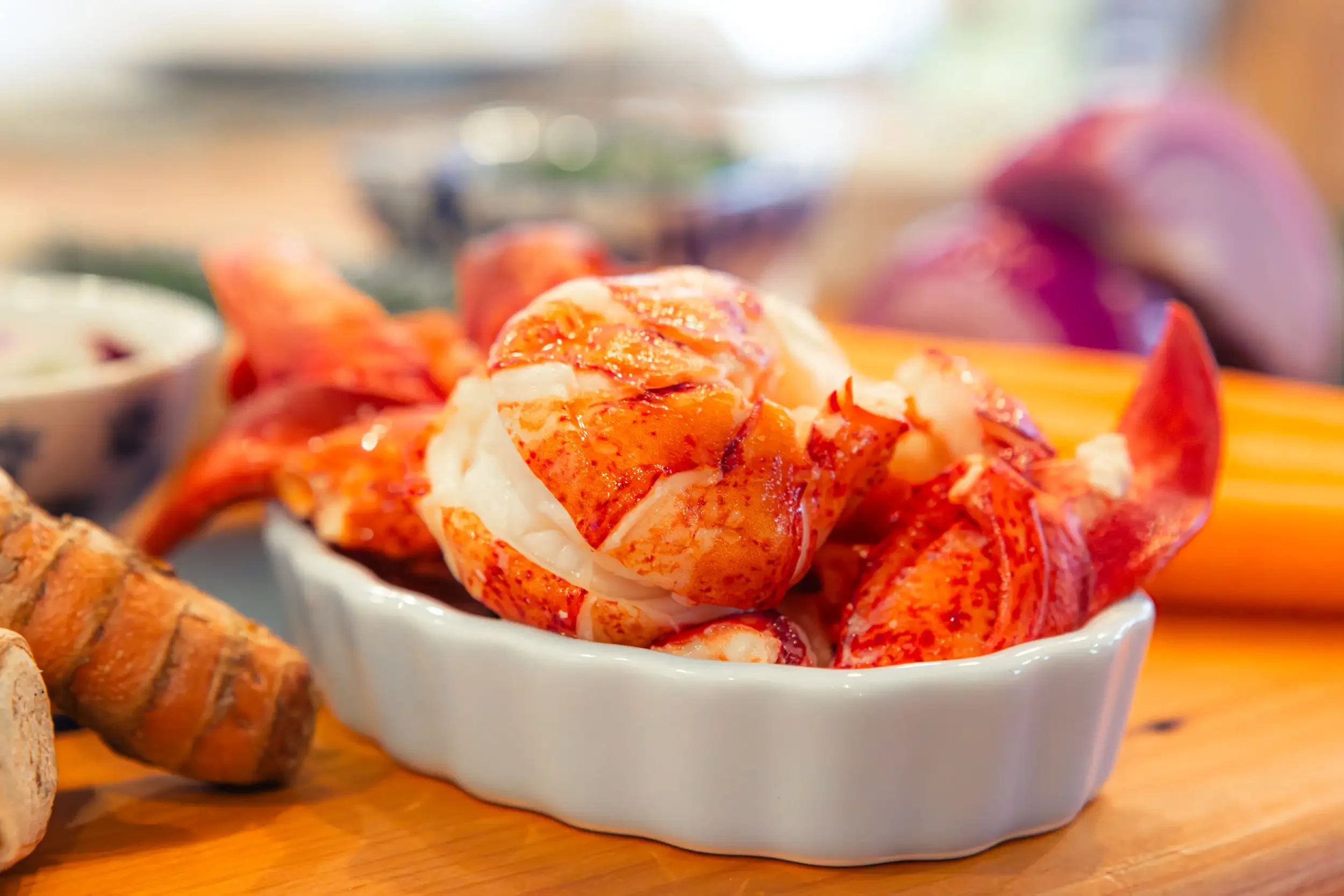 Close-up of cooked lobster tail served in a small white bowl, with other seafood dishes partially visible in the background.