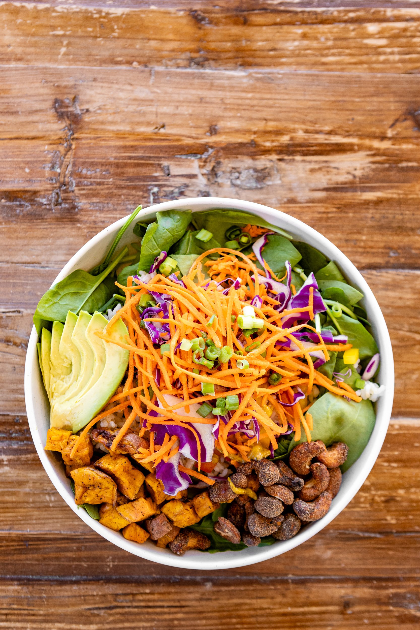 A bowl of salad with avocado, shredded carrots, purple cabbage, chopped green onions, roasted sweet potatoes, crumbled crispy toppings, and leafy greens, on a wooden surface.