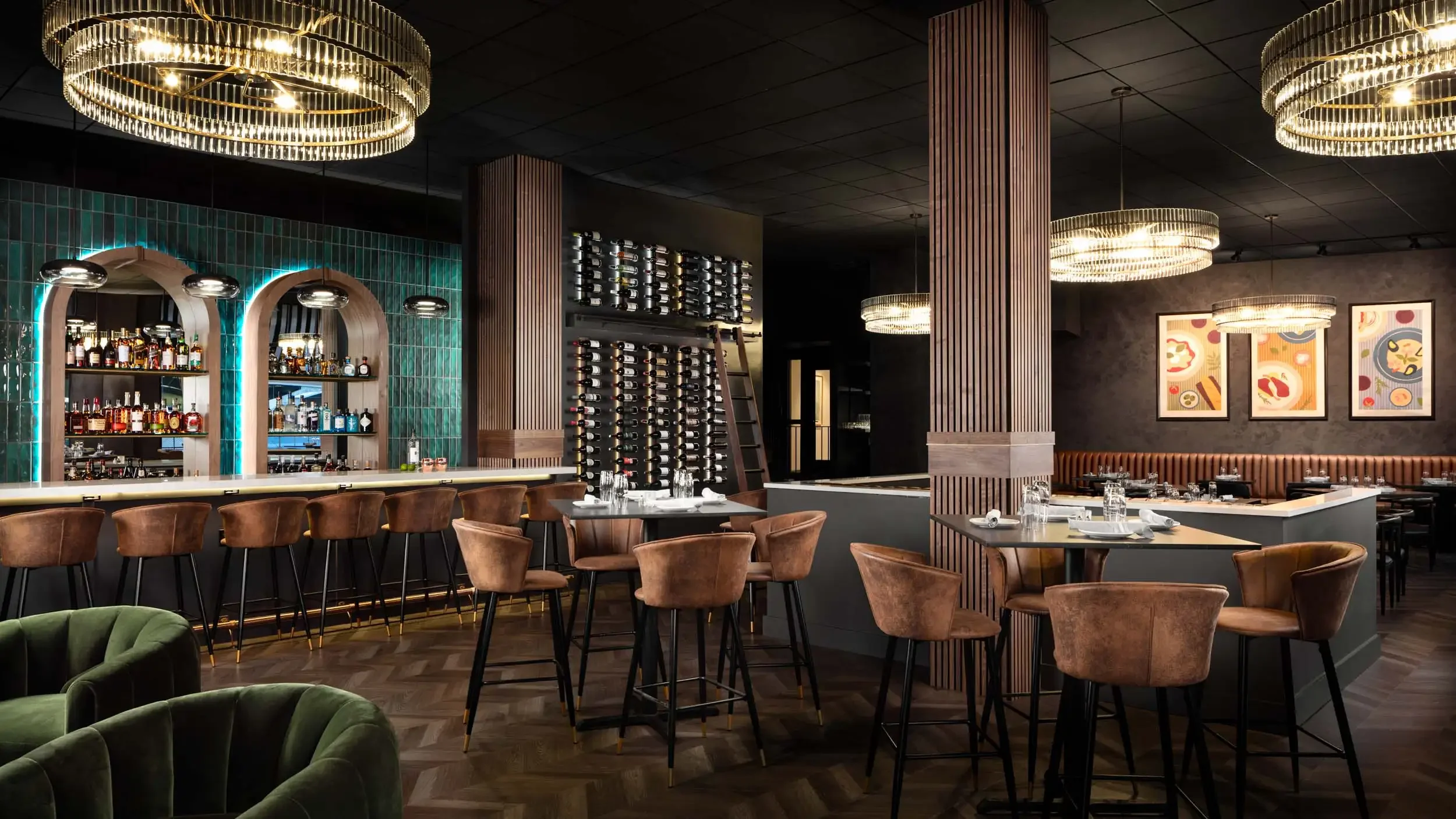 Interior of a modern upscale restaurant or bar with a dark ceiling, warm lighting, and wooden accents. There is a bar area with bottles on shelves, surrounded by brown chairs, and several tables set with glasses and napkins. Decorative framed artwork