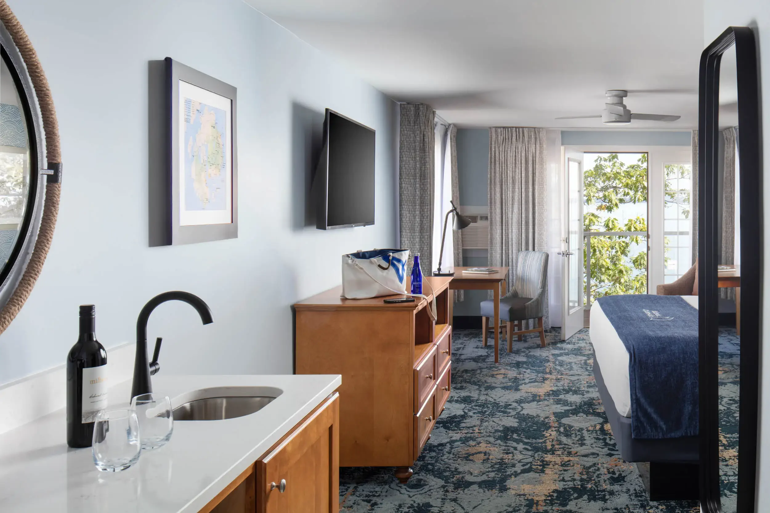 Interior of a hotel room with a small kitchenette, a dining table with chairs, a bed, a television on the wall, and an open balcony door with greenery outside.