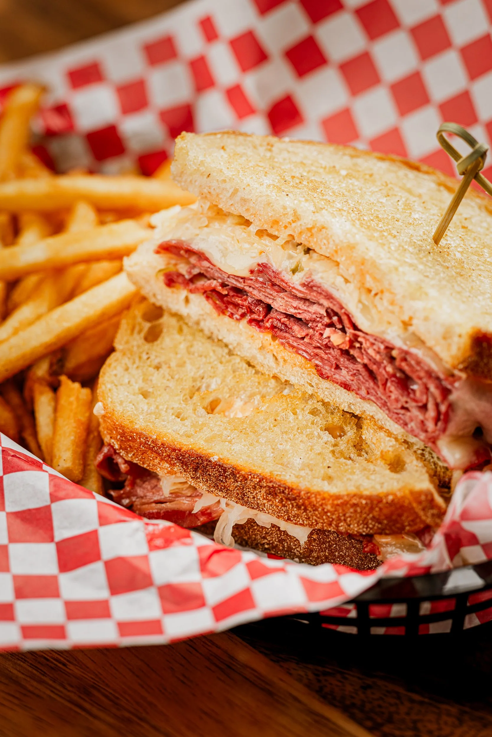 A reuben sandwich with sliced corned beef, sauerkraut, and melted cheese on toasted rye bread, served with French fries in a basket lined with red and white checkered paper.
