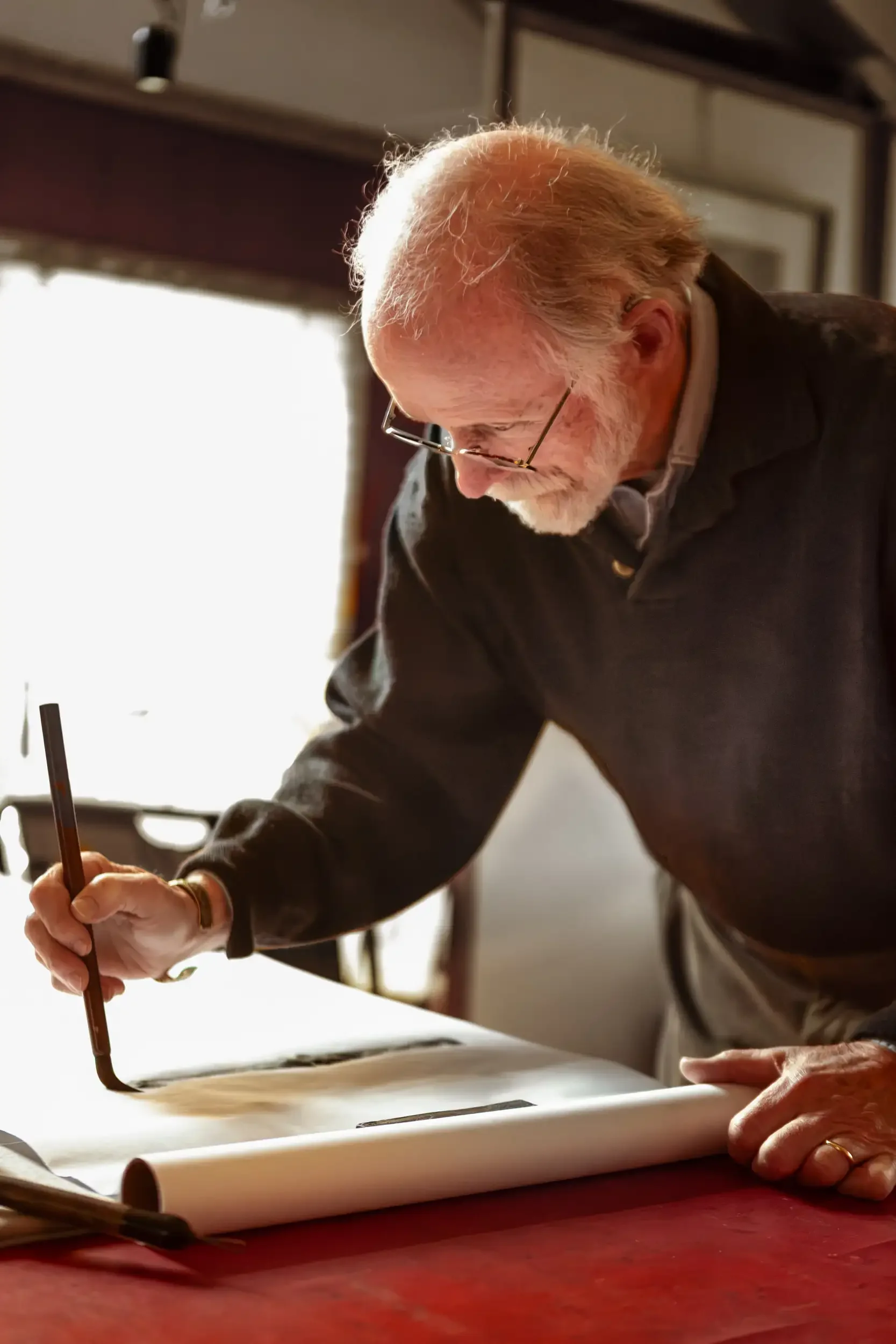 An elderly man with white hair and glasses is leaning over a table, writing or drawing on a large sheet of paper with a pencil. The background shows a window and a wooden interior.