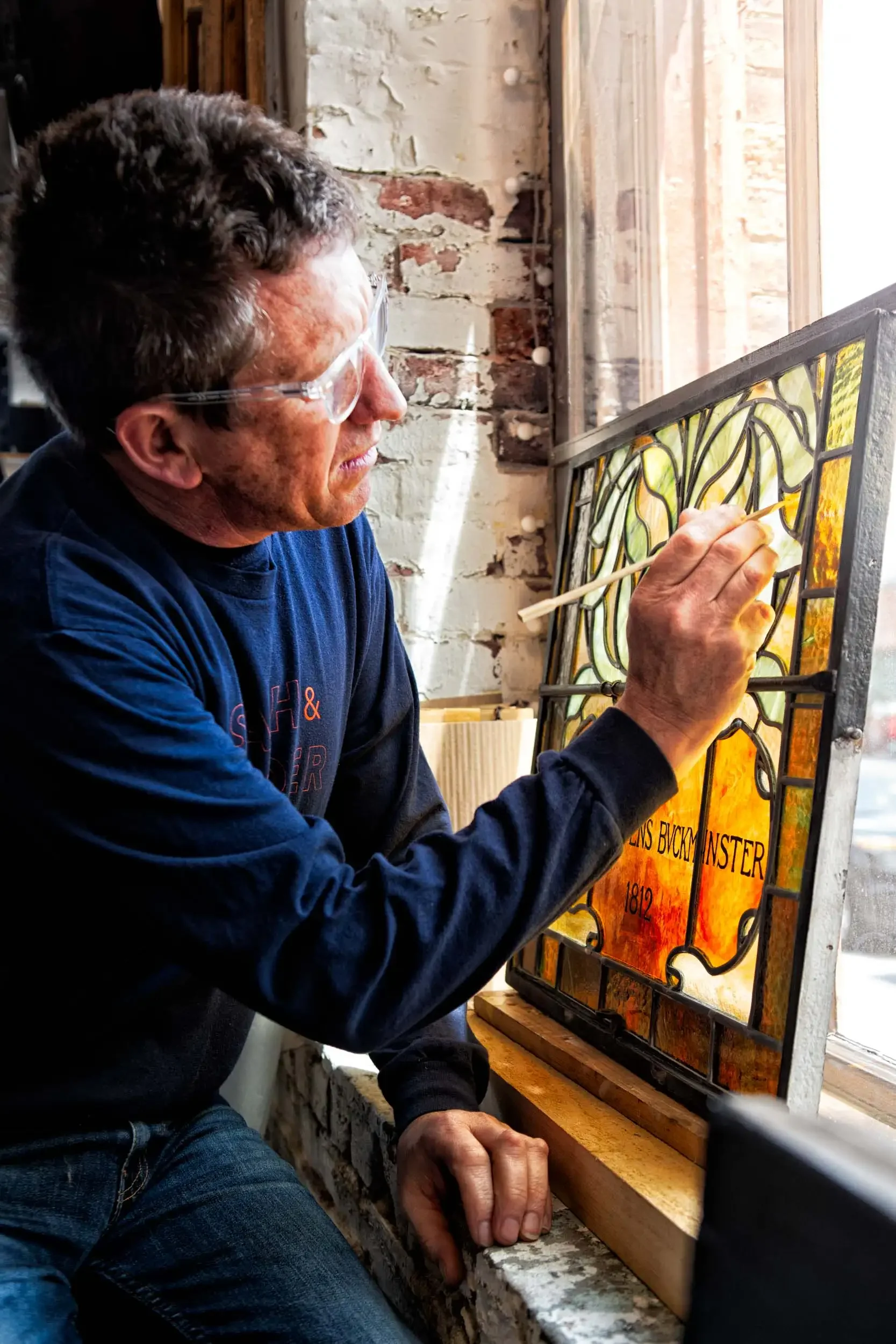 A man wearing safety glasses paints a stained glass window with a paintbrush, seated at a workbench in a rustic workshop with exposed brick walls.