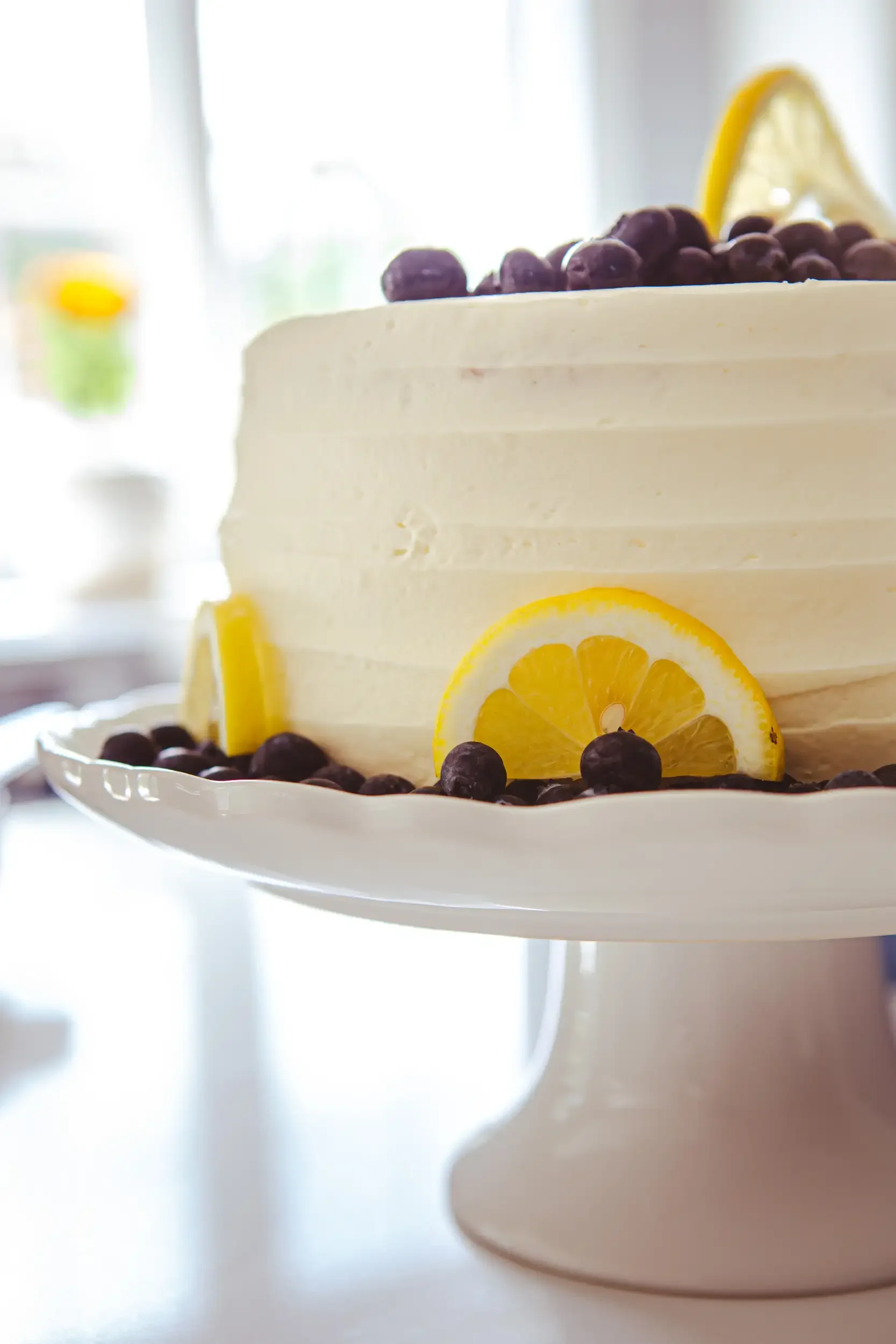 Close-up of a white frosted cake decorated with blueberries and lemon slices, presented on a white cake stand.