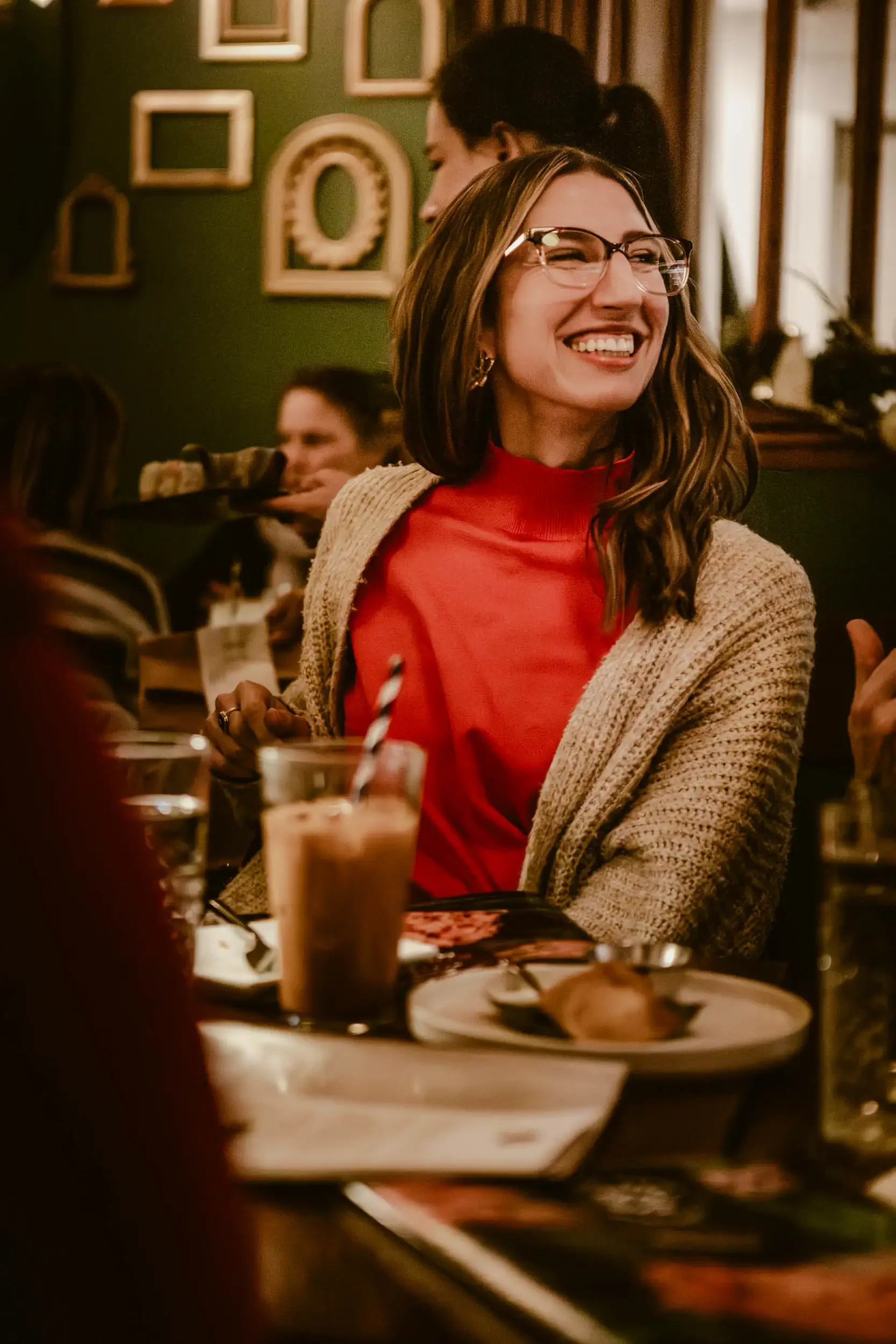 A woman with glasses and long hair smiling at a dining table in a cozy restaurant, with multiple glasses of beverages and a dessert on the table in the foreground, decorative framed pictures on the wall behind her.