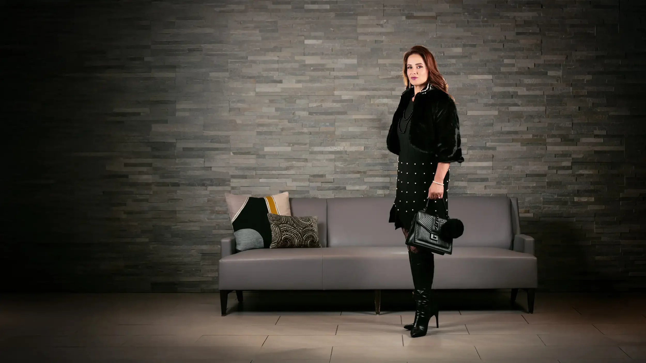 A woman in a black dress with pearl embellishments, black high-heeled boots, and a black fur jacket standing in a modern room with a gray stone wall, gray sofa, and decorative pillows.