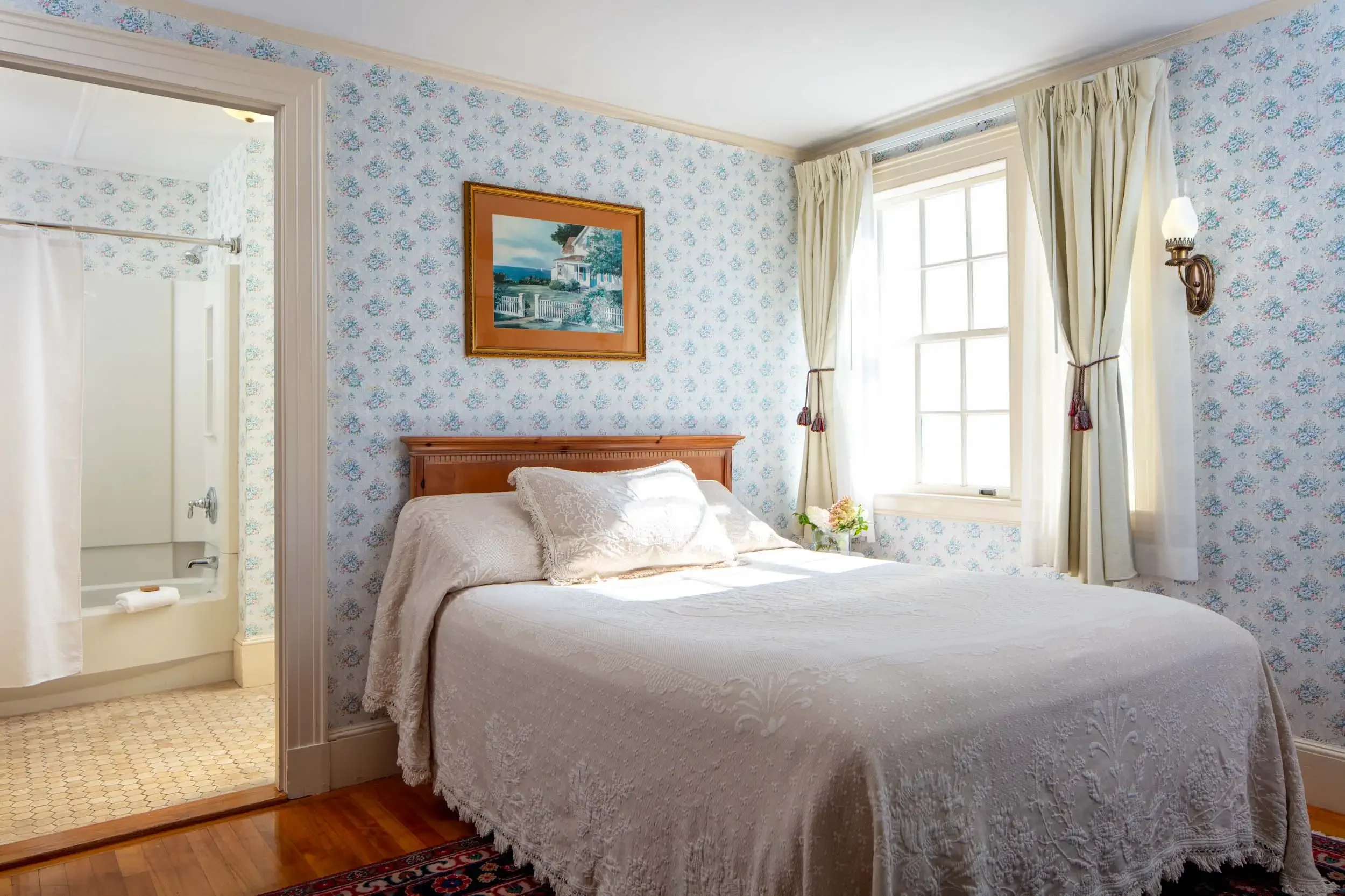 A cozy bedroom with a wooden bed frame, white bedding, floral wallpaper, large window with cream curtains, and a framed painting of a house outside. A small flower arrangement sits on the nightstand.
