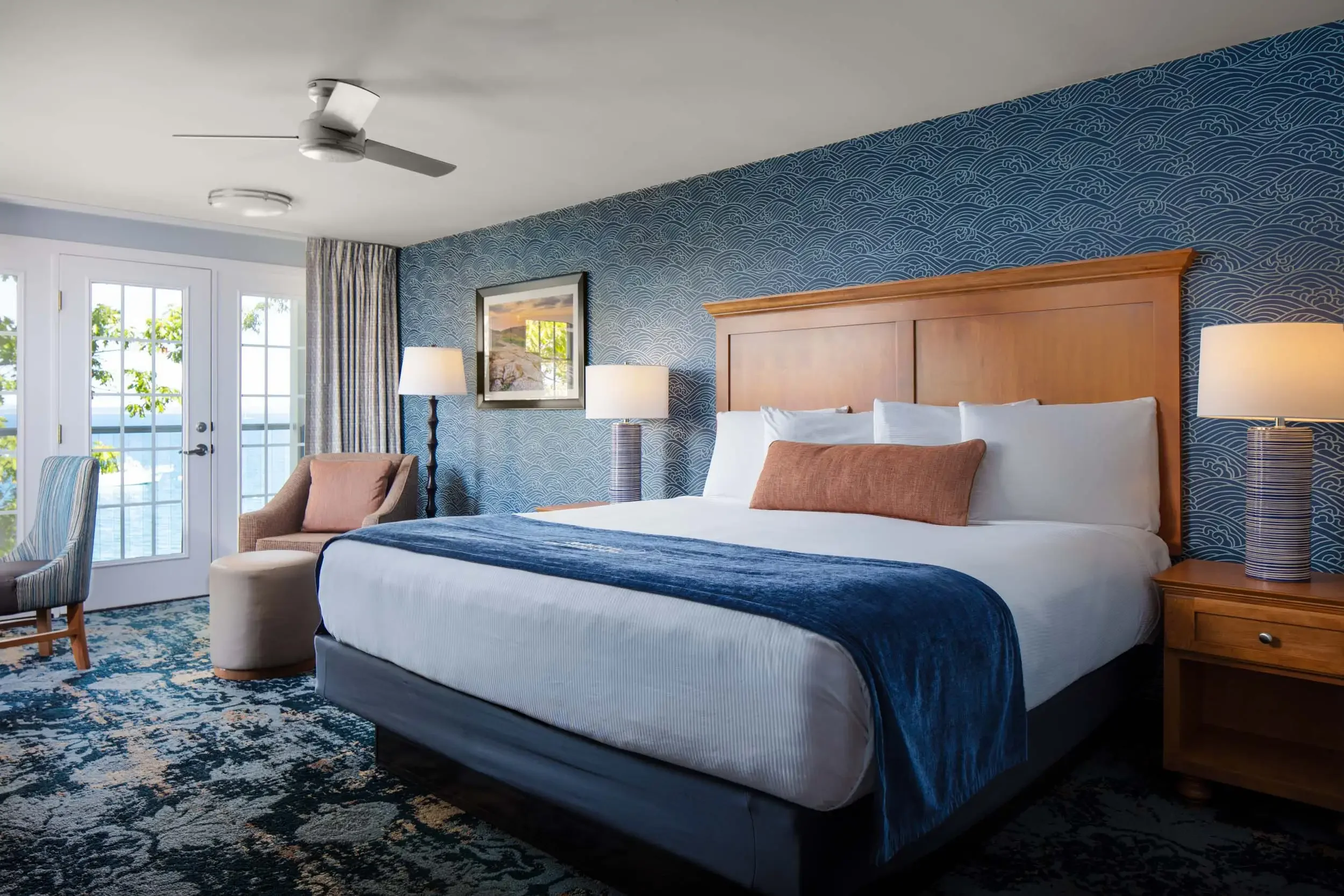 A hotel room featuring a large bed with white linens, pillows, and a blue throw blanket. The room has a wooden headboard, nightstands with lamps, a patterned blue wallpaper, a framed picture, a chair, a round ottoman, and glass doors leading outside 