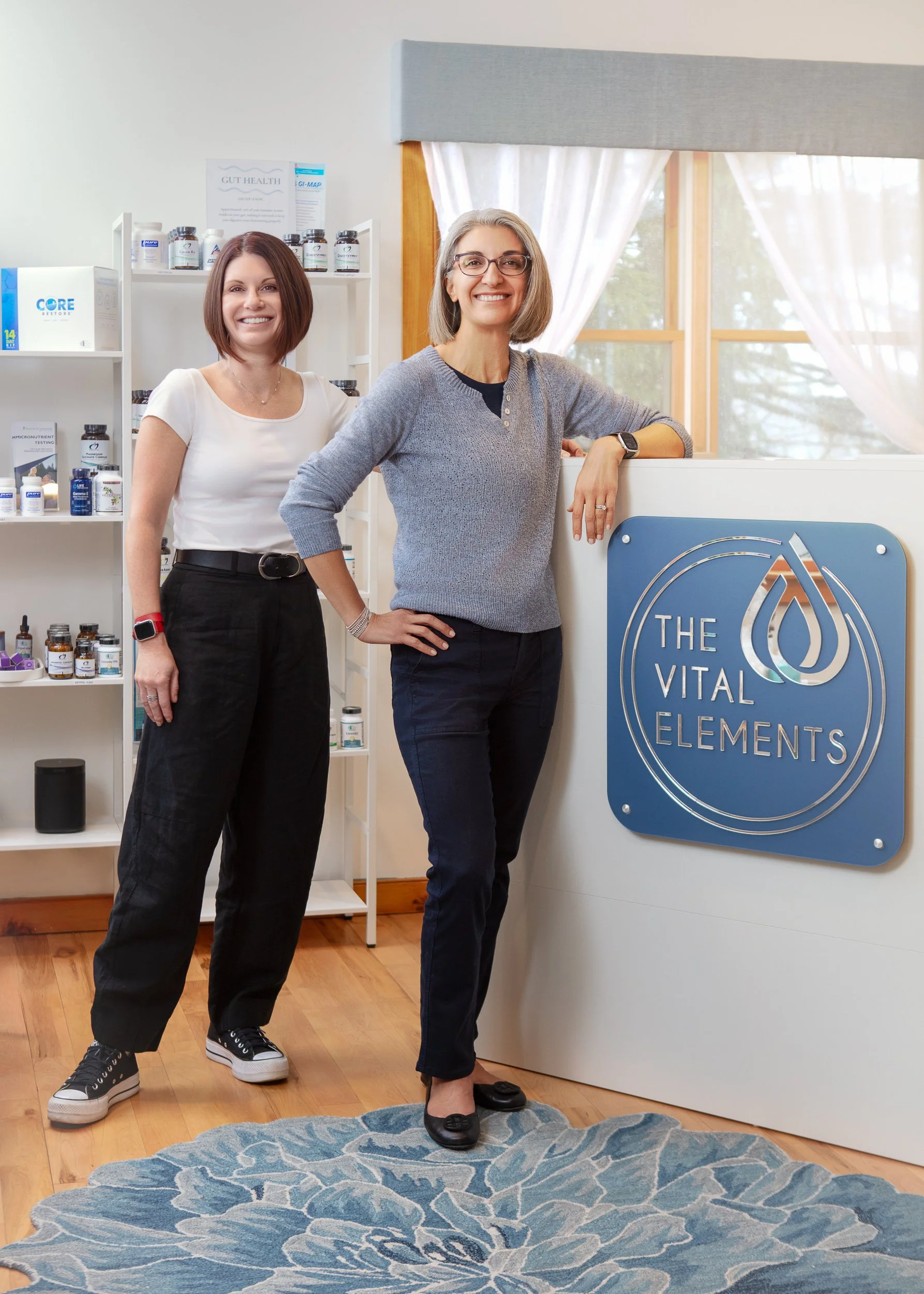 Two women standing inside a wellness or health clinic, near a reception desk with a sign that reads 'The Vital Elements'. One woman is wearing a gray sweater and glasses, and the other is in a white t-shirt with black pants. Shelves with health suppl