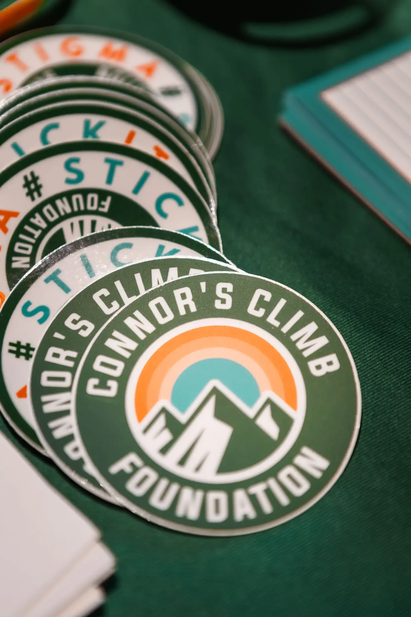 Close-up of circular stickers with the text '#STICK' and 'YOUTH FOUNDATION' and logo of mountains and a sun on a green surface, with a deck of cards nearby.