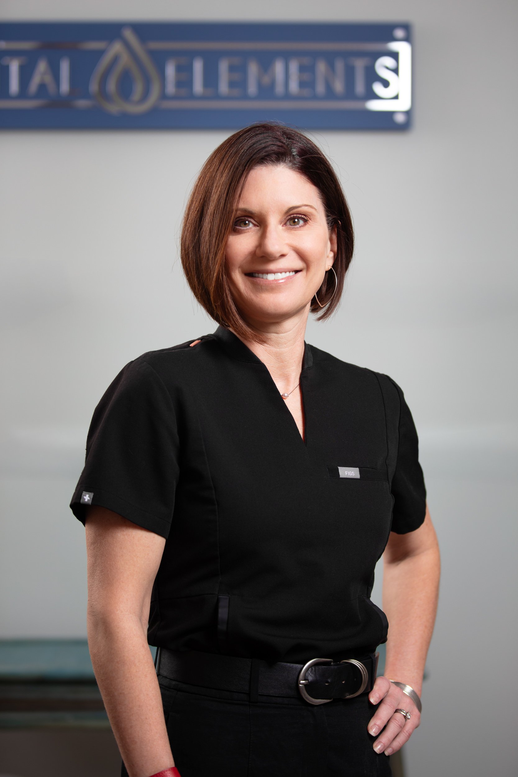 A woman with shoulder-length brown hair, wearing a black medical uniform, standing with her hand on her hip and smiling in front of a sign that says 'Total Elements'.