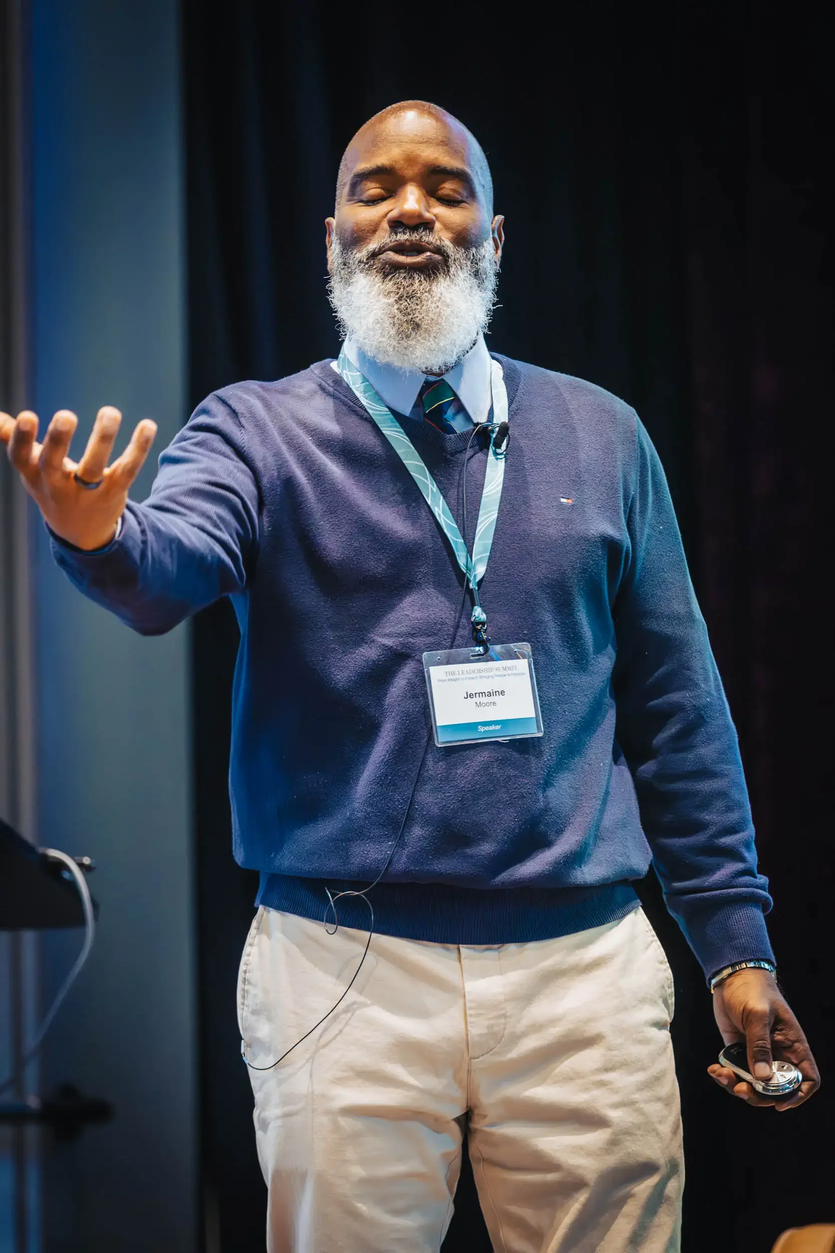 A man with a gray beard, wearing a blue sweater, white pants, and a lanyard with a badge, is standing on stage giving a presentation. He is holding a remote control in his right hand and has a microphone attached to his sweater.