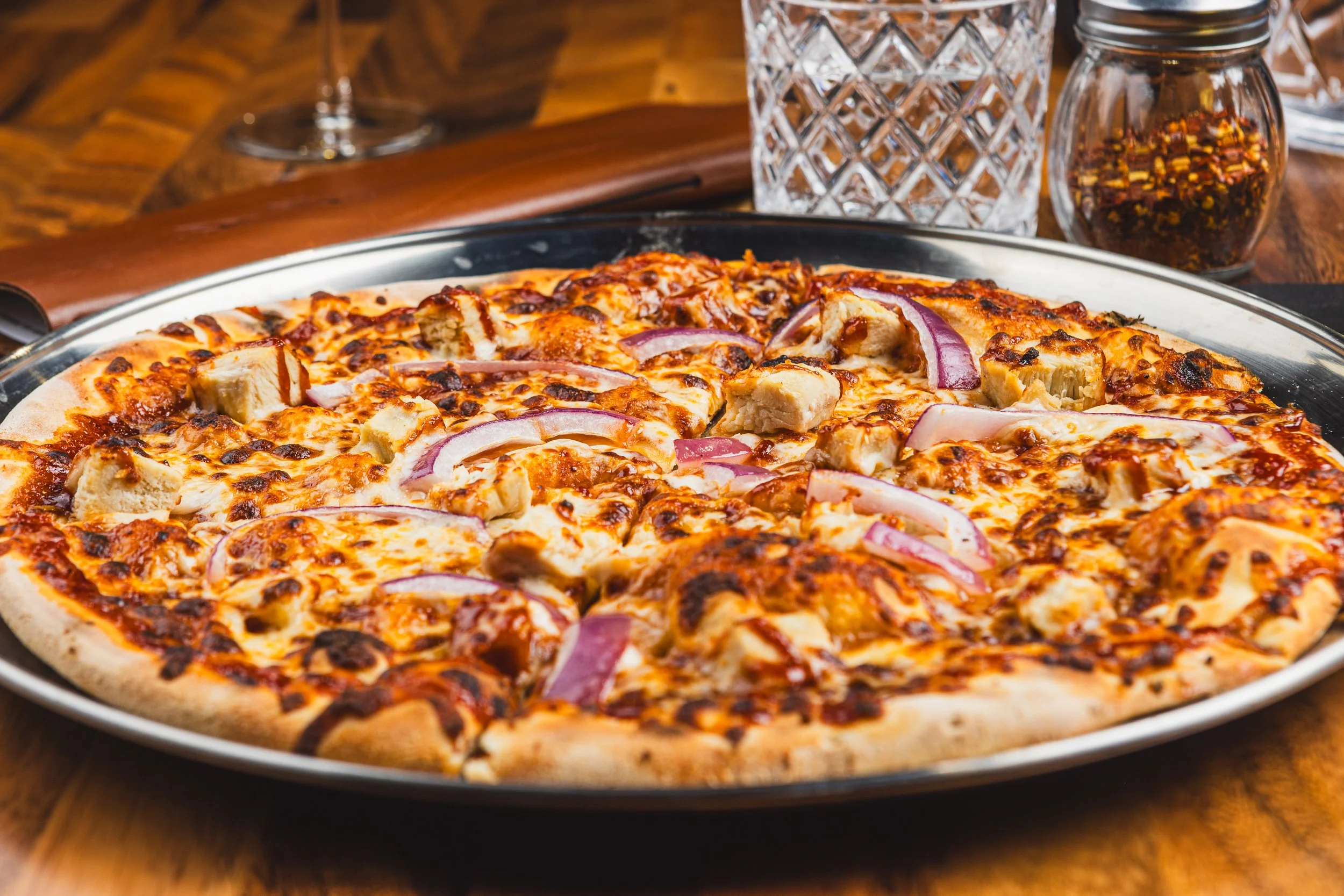 A pizza topped with chicken chunks, red onions, and cheese on a silver tray. Glasses and condiments are in the background.