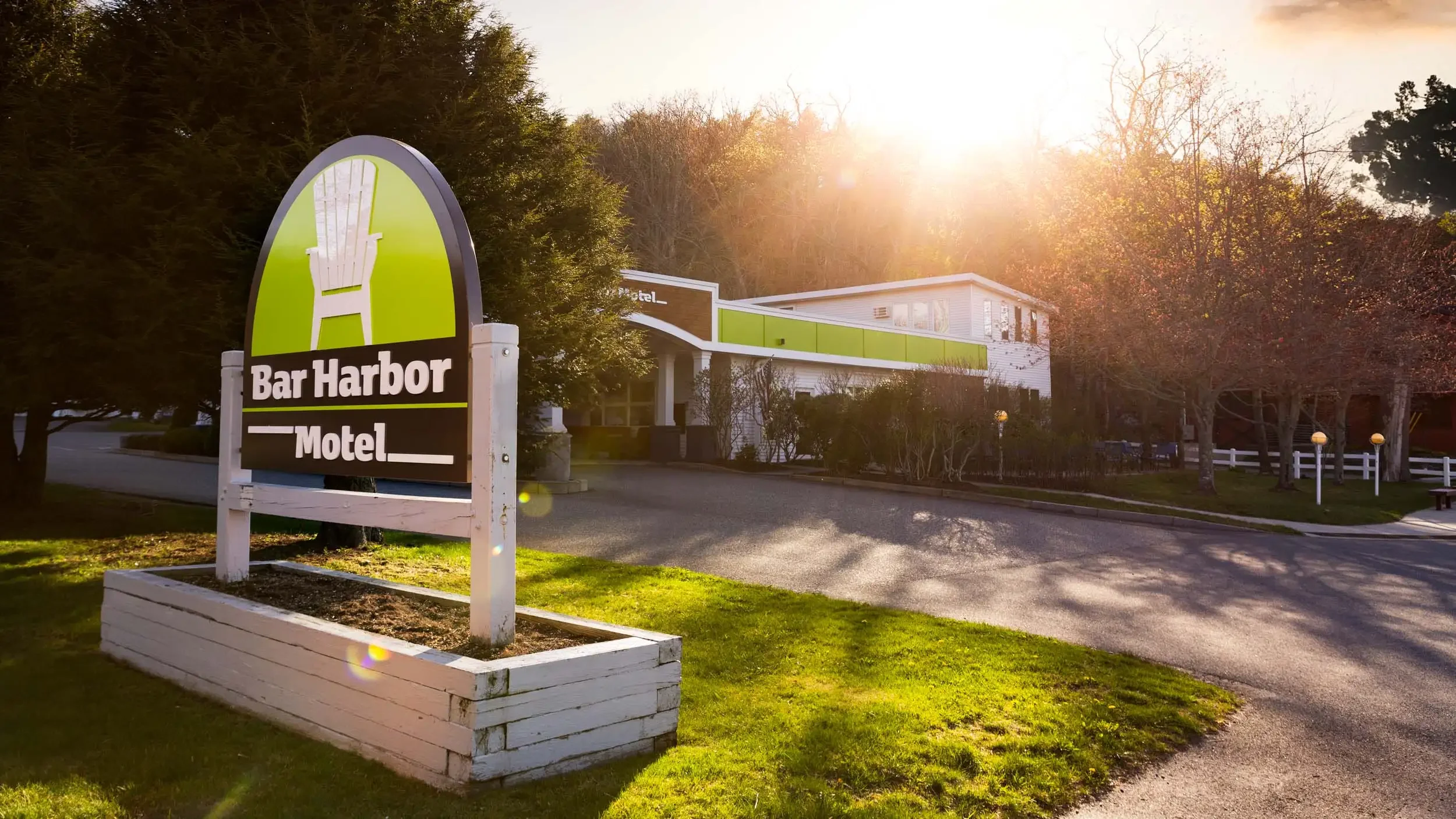A sign for Bar Harbor Motel featuring a white Adirondack chair on a green background, with the motel building in the background during sunset, surrounded by trees and a well-maintained lawn.