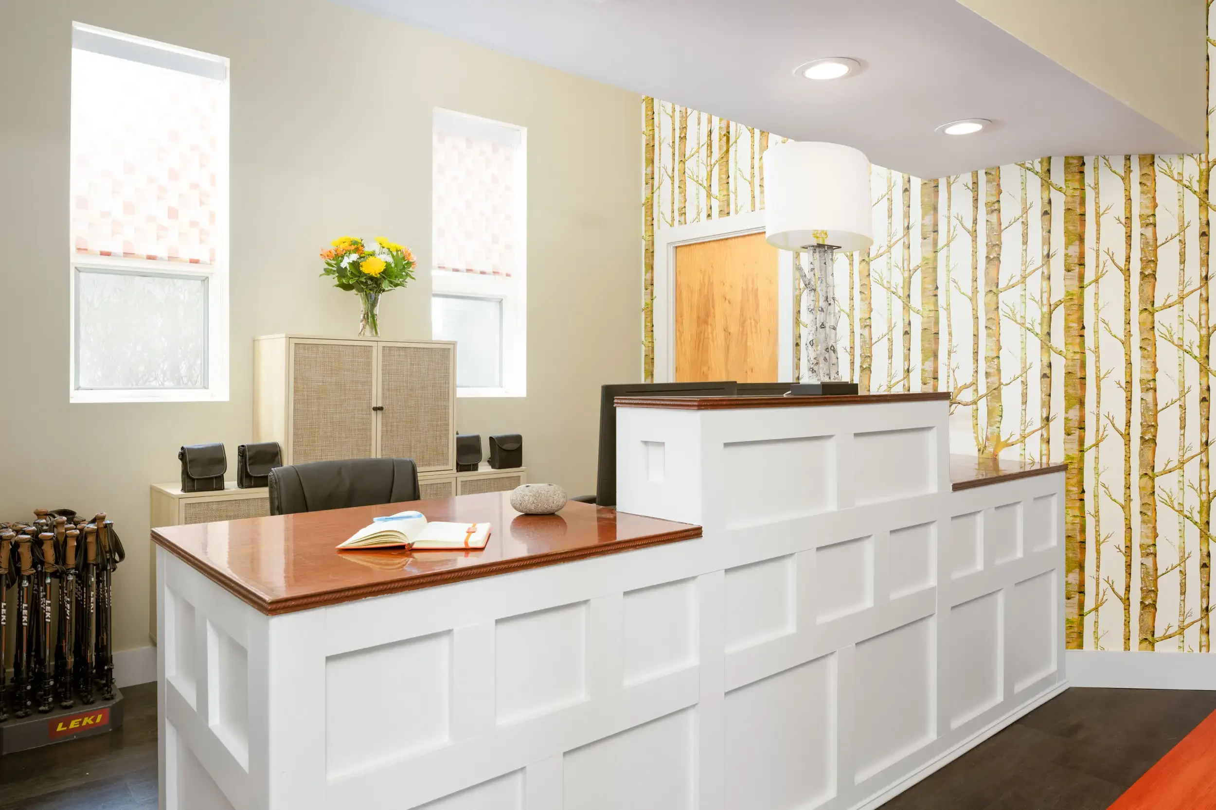 Reception area with a white checkered counter, a wooden desk, a book, a stone, a vase with yellow flowers, and windows with roller shades, and a wallpapered accent wall with a birch tree pattern.
