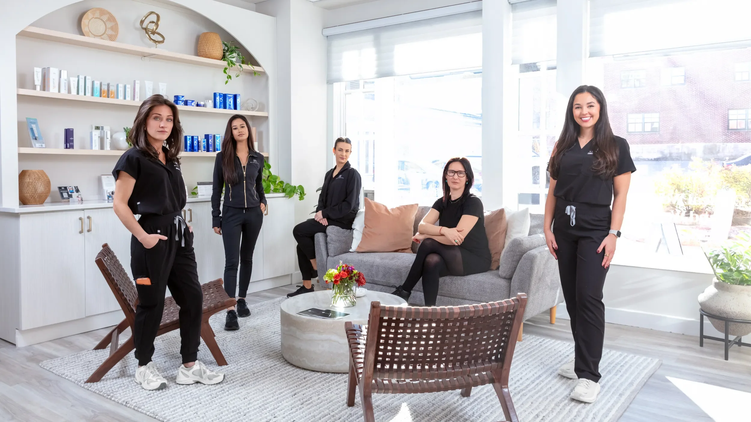 Five women inside a modern, well-lit living room with large windows, a sofa, a coffee table with a vase of flowers, and decorative items on shelves, posing for a group photo.