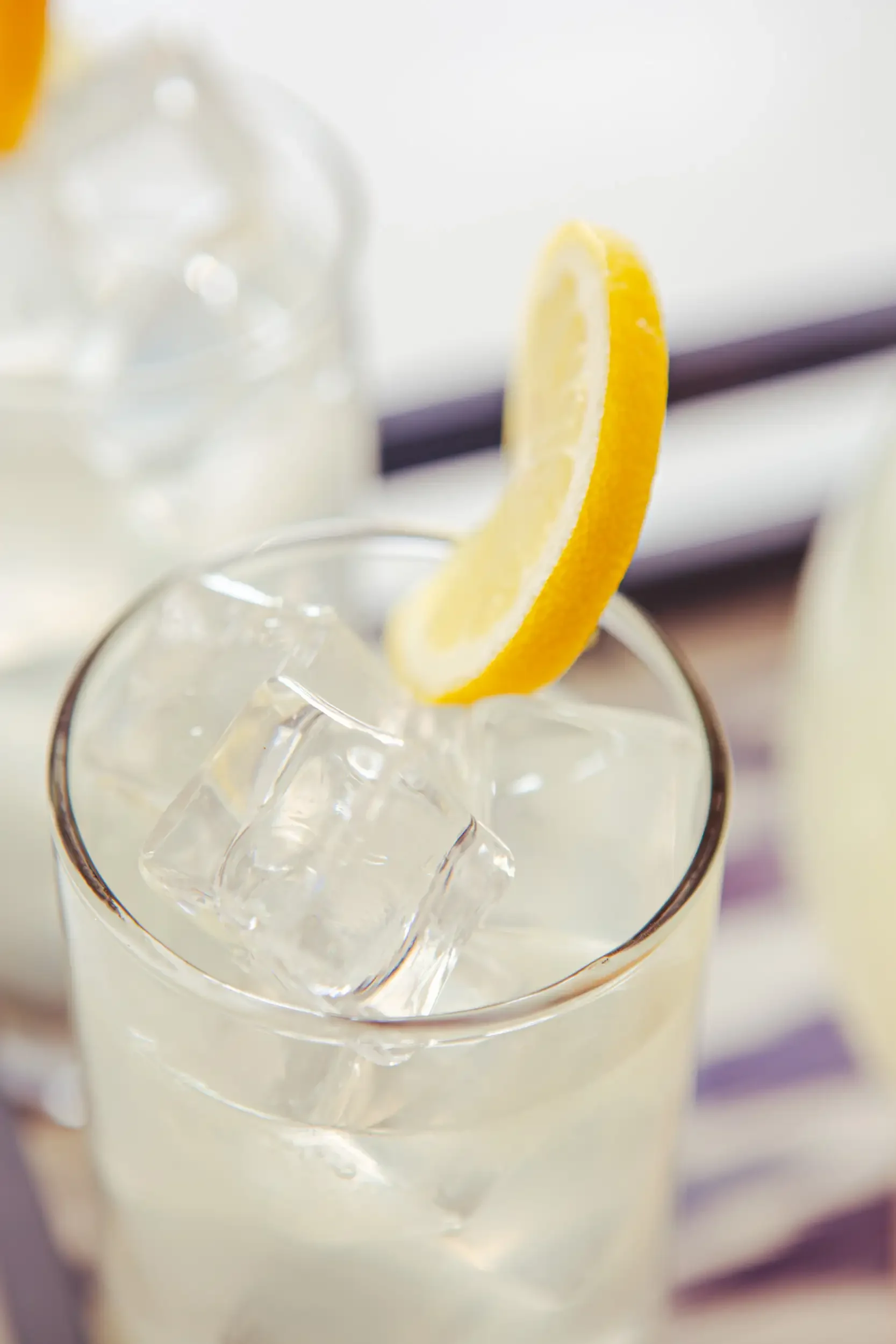 A close-up of a glass with ice cubes and a lemon wedge garnish, with a blurred background.