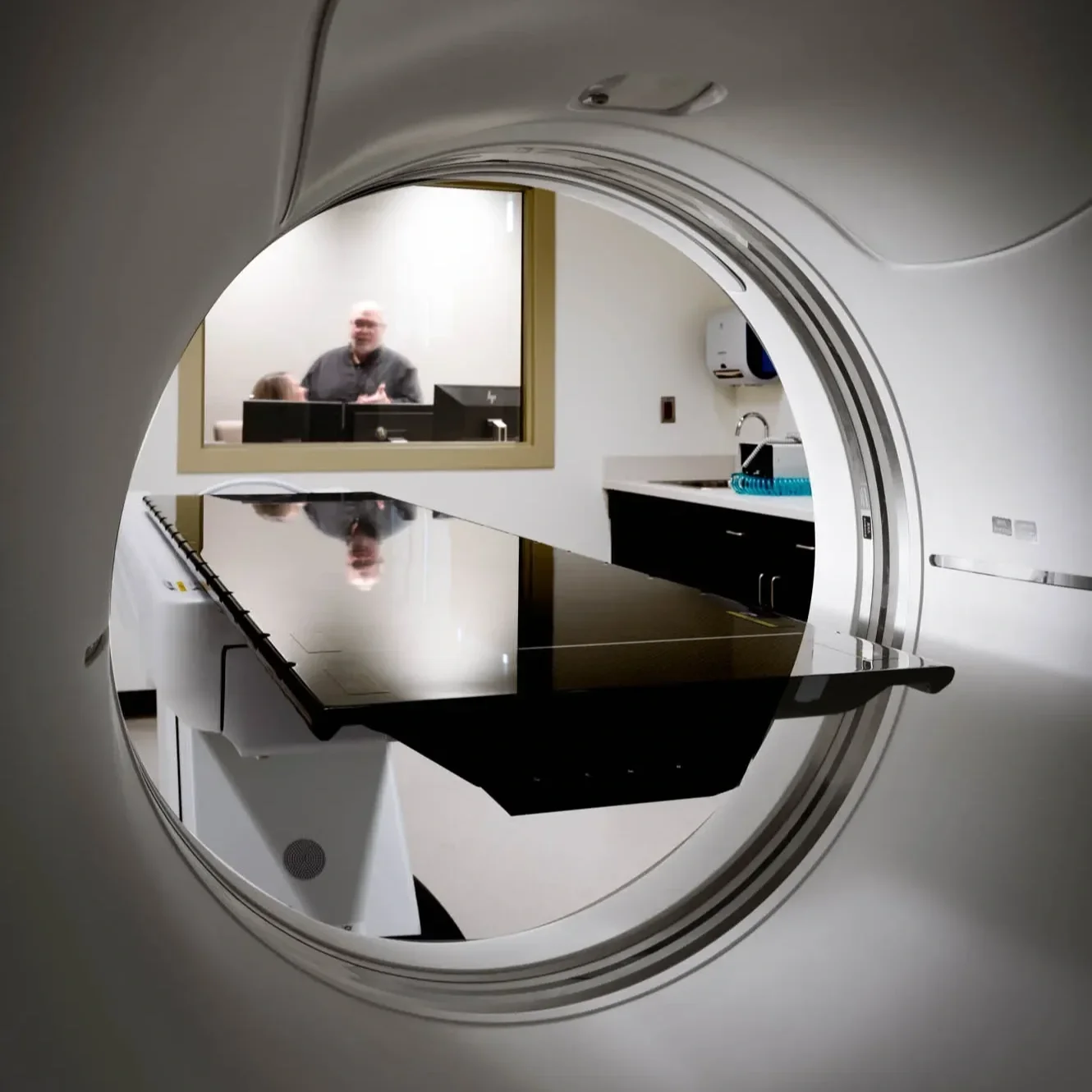 MRI machine with a reflective black surface, interior of a medical room, and a technician and patient visible through the viewing window.