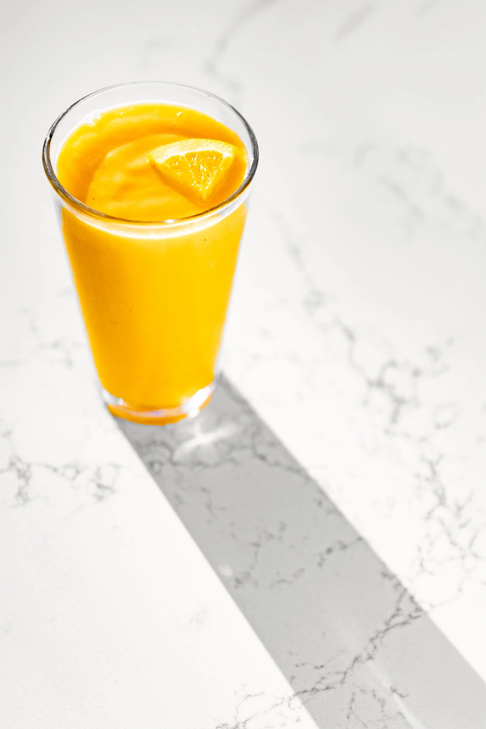 A tall glass of orange juice with a slice of orange on top, placed on a white marble surface with shadows.