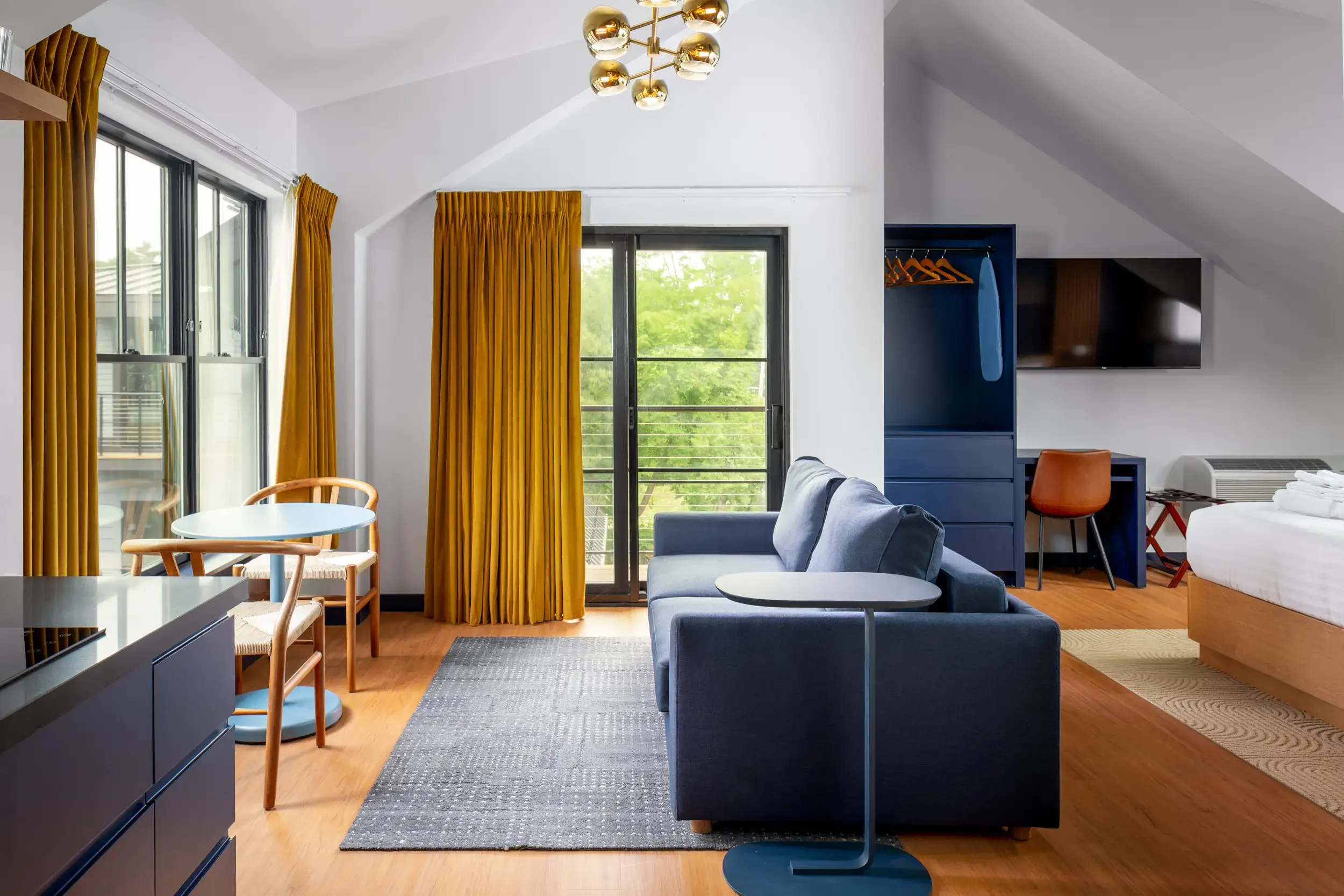 A modern living room with large windows and yellow curtains, a blue sofa with matching armchair, a round white coffee table, a small blue dining table with chairs, a wall-mounted television, and a small closet with hanging clothes. There's also a bed