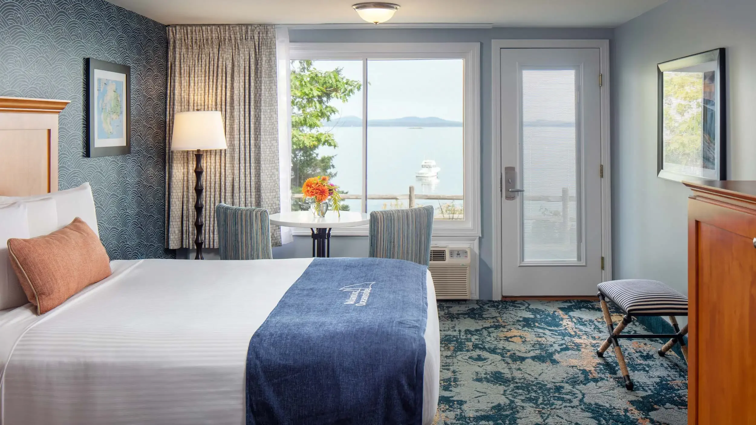 Hotel room with a bed, a window with a view of water and boats, a table with a flower arrangement, and patterned wallpaper.