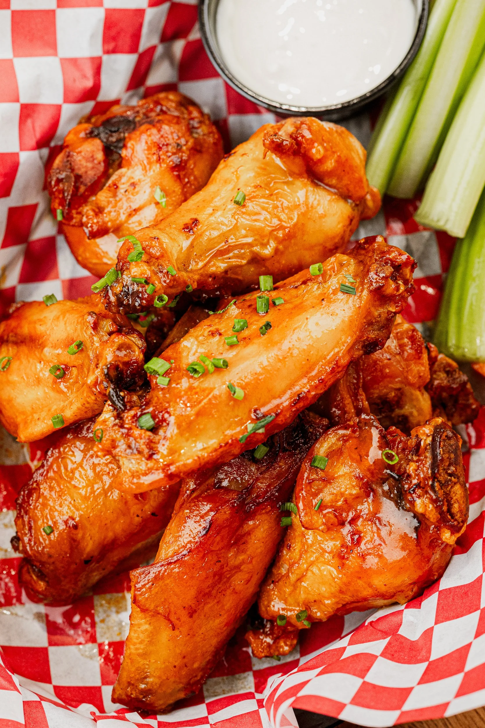 Close-up of cooked chicken wings garnished with chopped green onions, served with celery sticks, a small cup of ranch dressing, and a cup of dipping sauce, on checkered red and white paper.