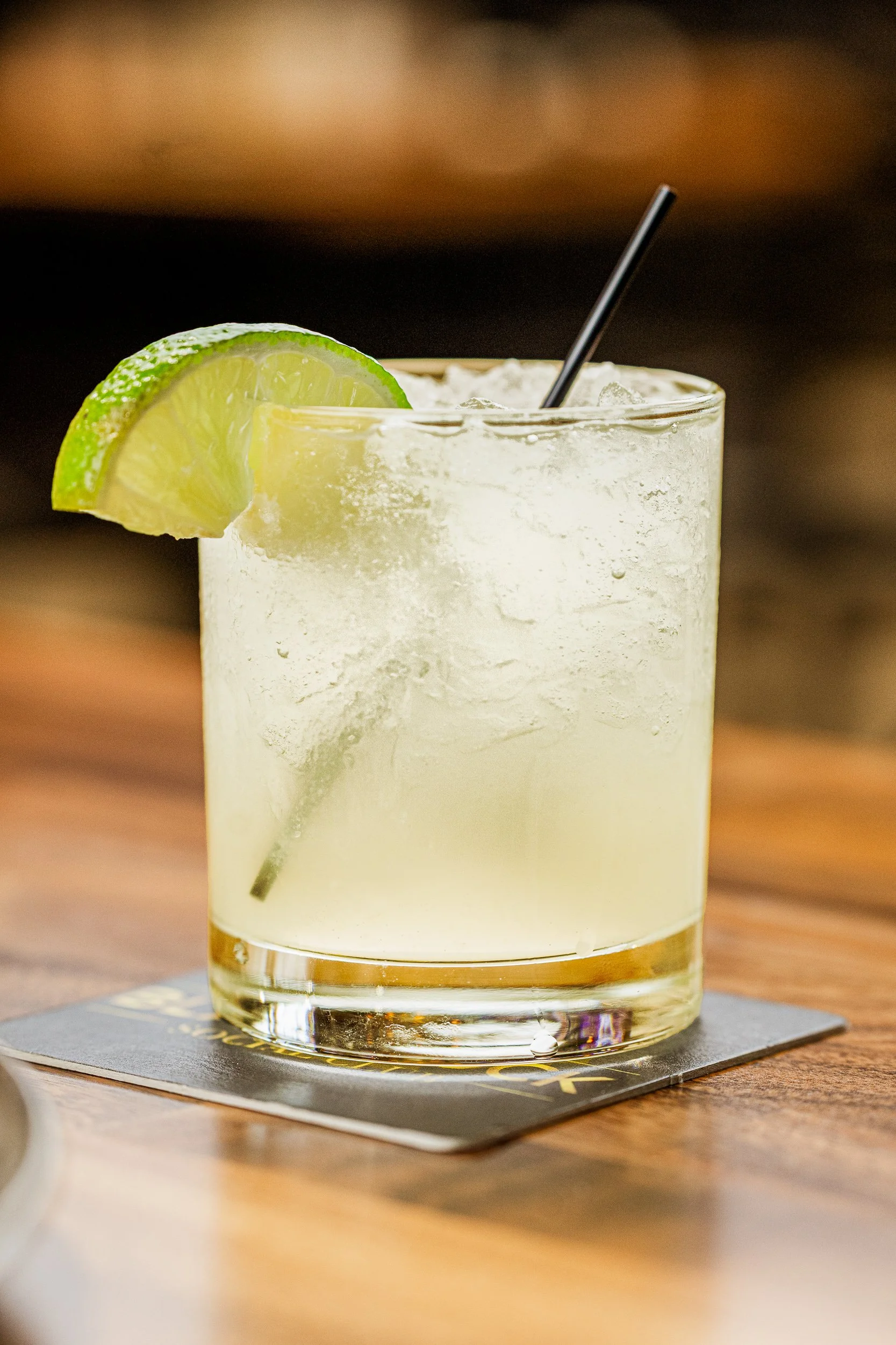 A glass of pale yellow cocktail with lime wedge garnish and a black straw, placed on a coaster on a wooden surface.