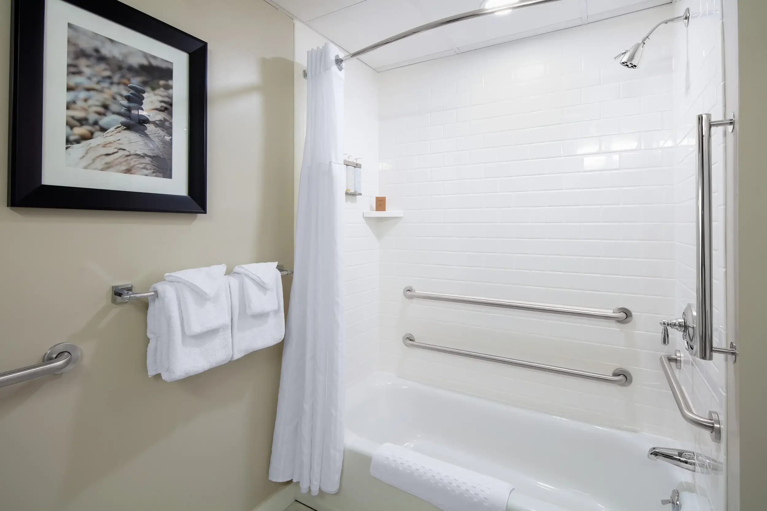 A bathroom with a white bathtub and shower, a shower head, and safety grab bars. There are white towels hanging on a silver towel rack, and a framed picture of stacked stones on the wall.