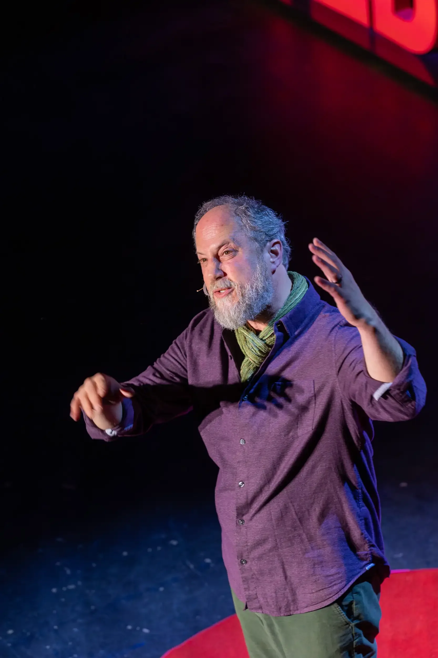 A man with a beard and receding hairline speaks on stage, gesturing with his right hand. He wears a maroon button-up shirt and a green scarf. The background is dark with red and purple lighting.