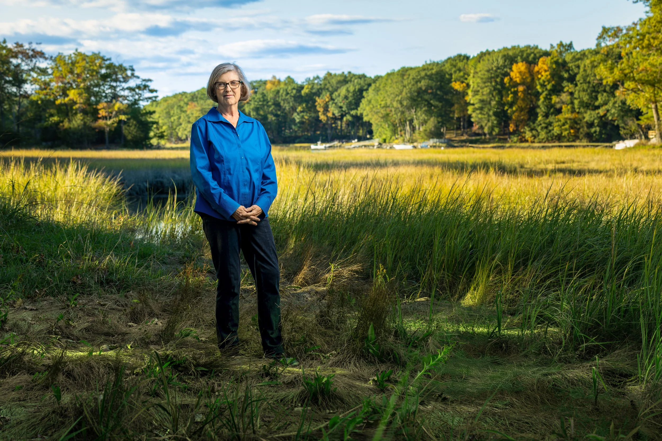 A woman with gray hair wearing glasses, a blue shirt, and black pants standing outdoors near a marshy area with tall grass and a wooded background.