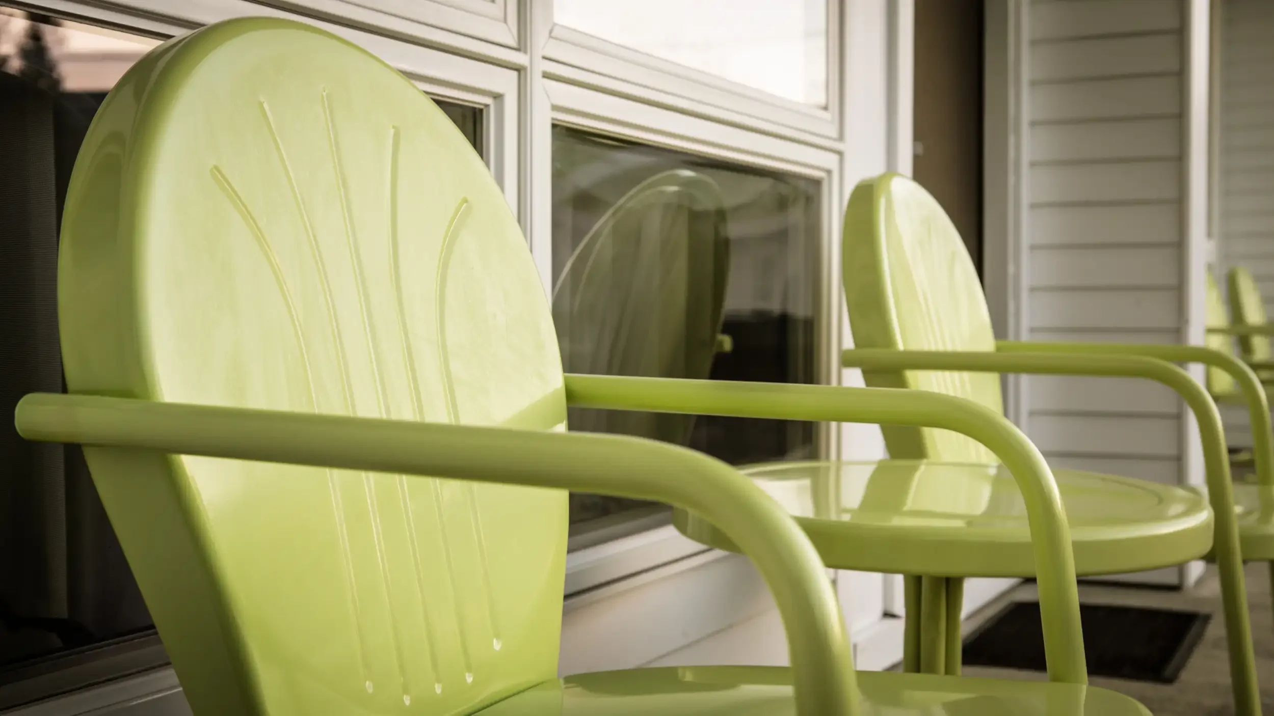 Close-up of lime green outdoor chairs on a porch or balcony with a window and siding in the background.