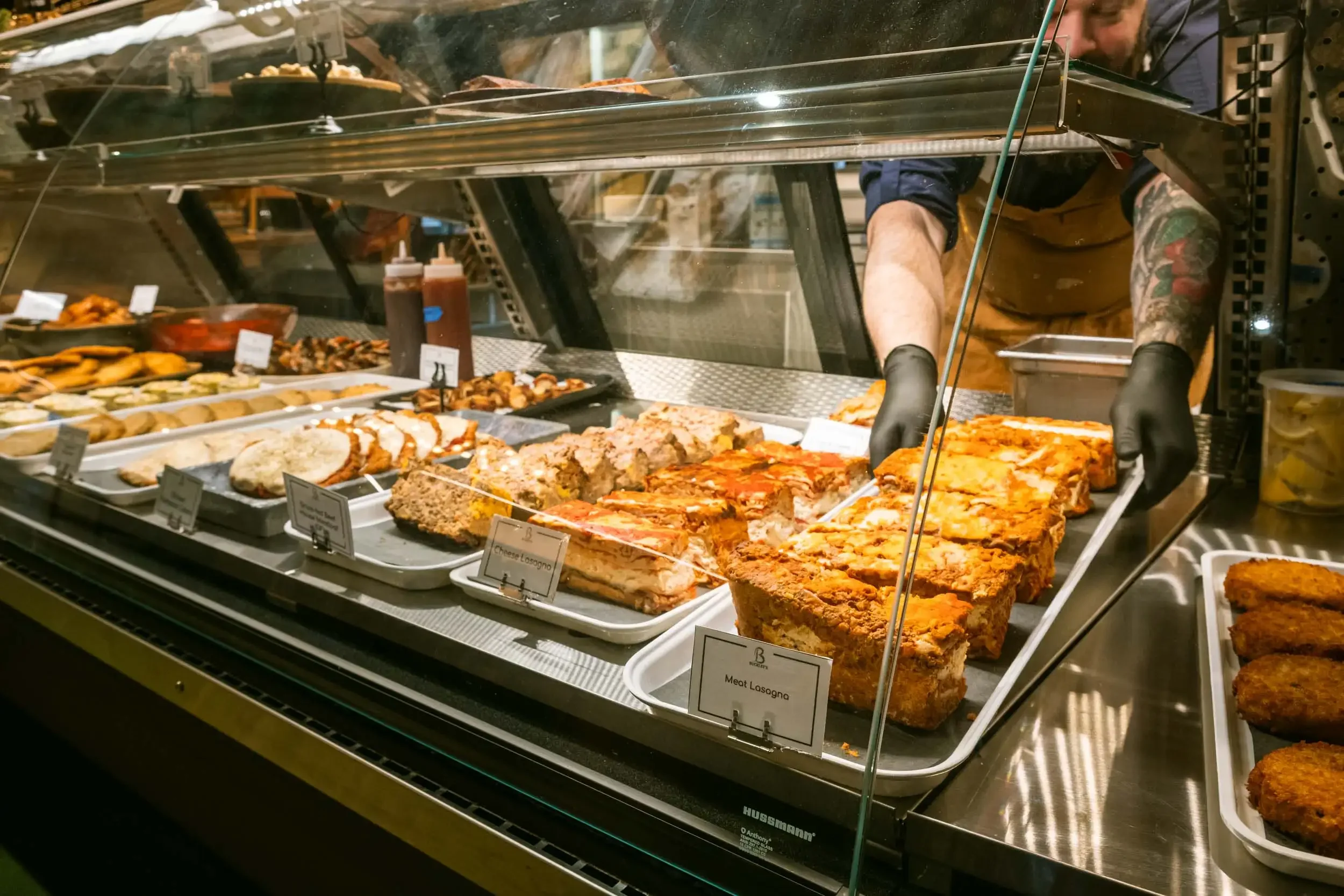 Display case with various slices of pizza and baked dishes at a restaurant or deli. A worker is seen behind the glass, wearing gloves and a tattooed arm.