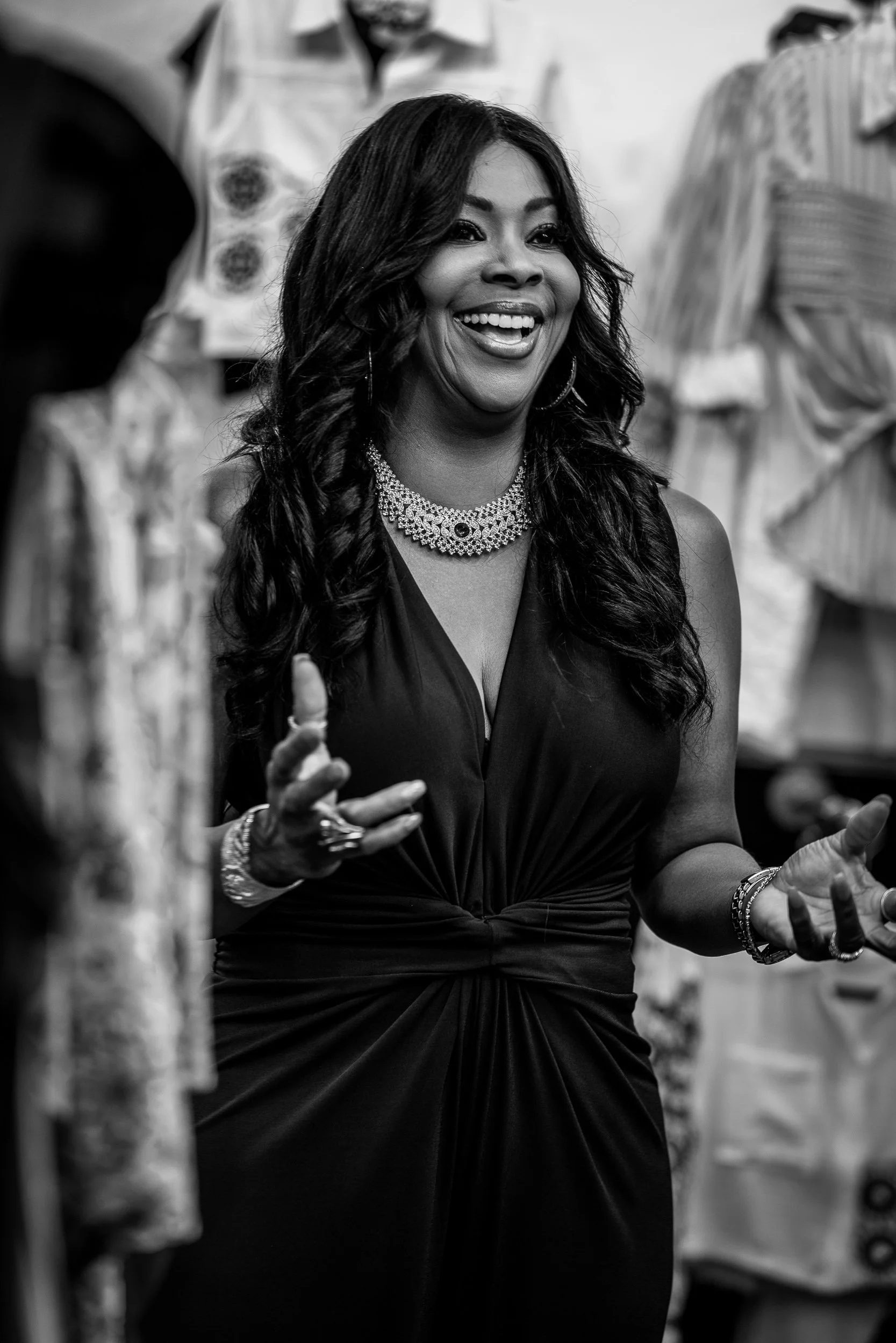 Smiling woman with long dark hair, wearing jewelry and a sleeveless dress, talking in a social setting, with blurred people and clothing in the background.