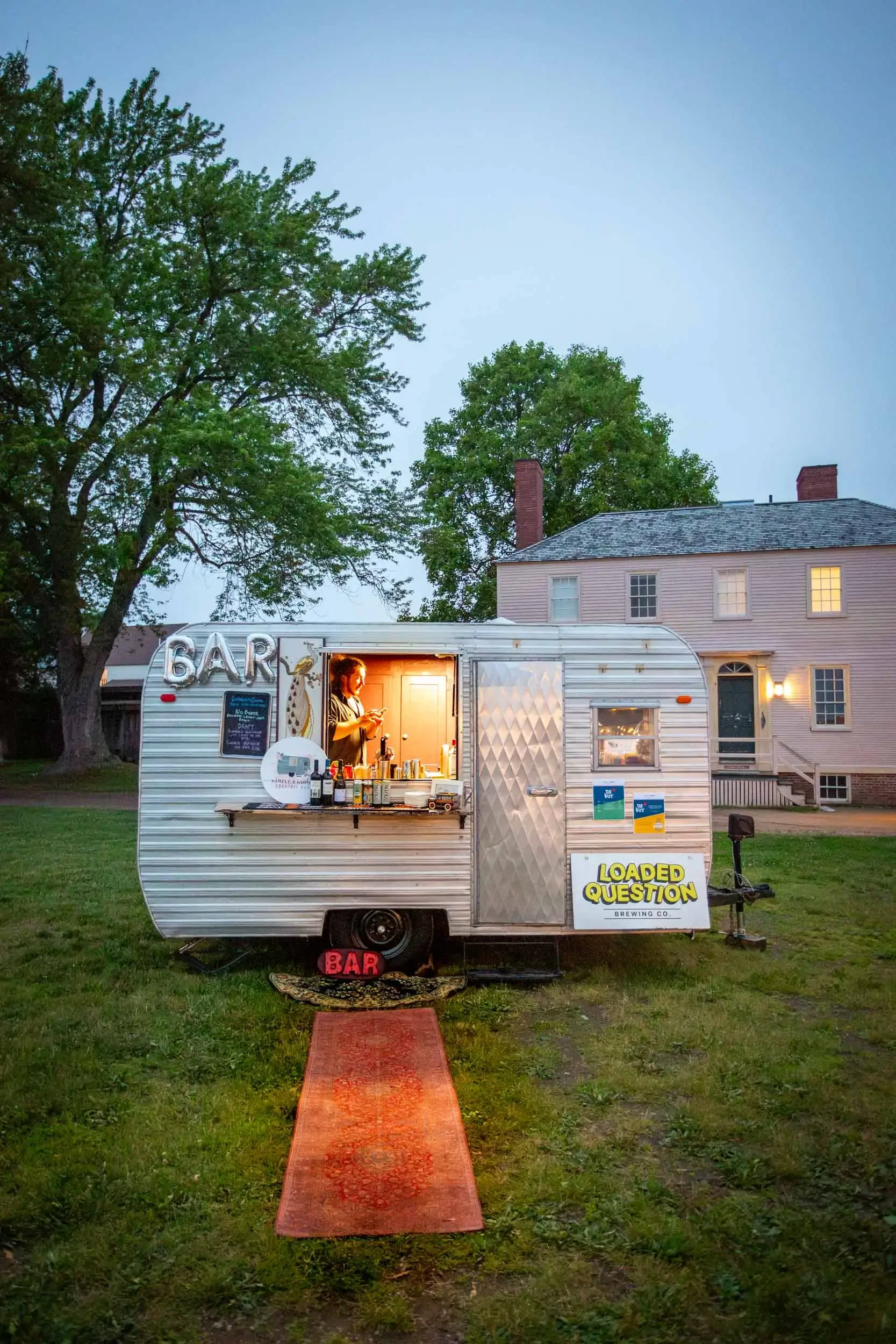 A vintage camper trailer serving as a bar, with large illuminated 'BAR' letters and a chalkboard menu outside. A woman inside is preparing drinks. The scene is set outdoors in a grassy area with a red carpet leading to the trailer and a house with li