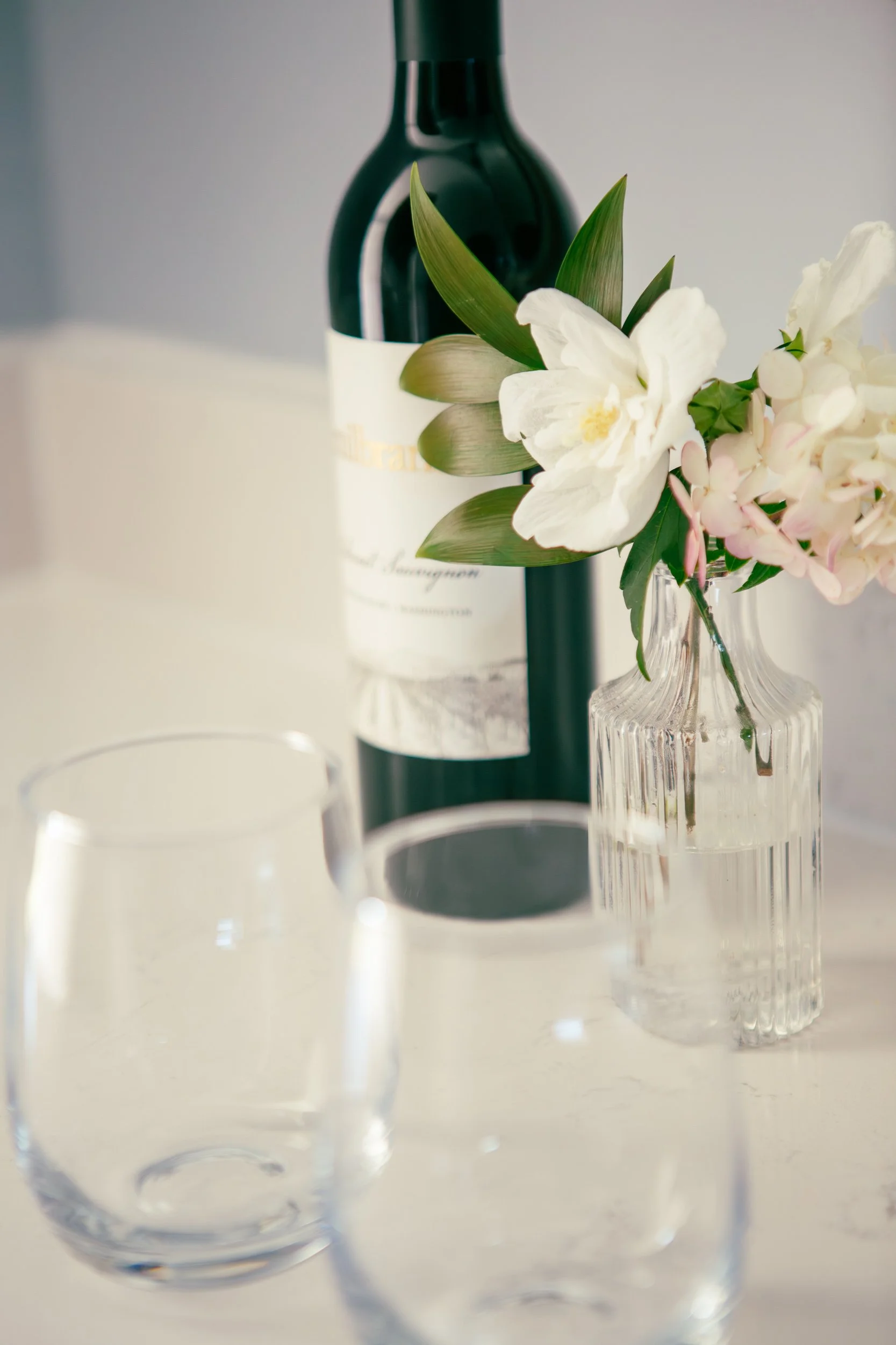 A bottle of white wine in the background with a white label, a clear glass vase with white and pink flowers, and two empty clear drinking glasses on a light-colored surface.