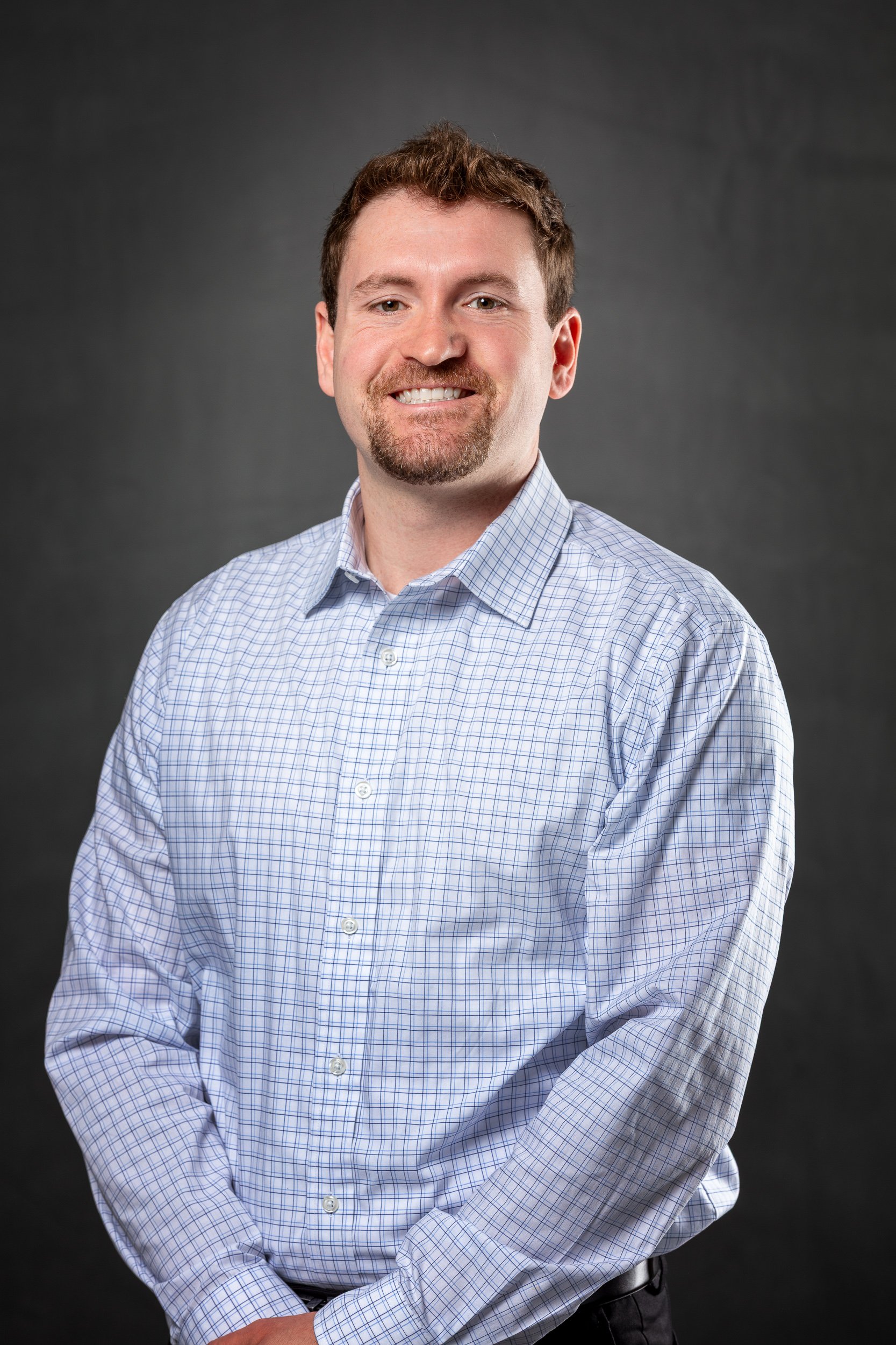 A professional headshot of a smiling man with short brown hair and a beard, wearing a light blue checkered dress shirt, against a dark gray background.